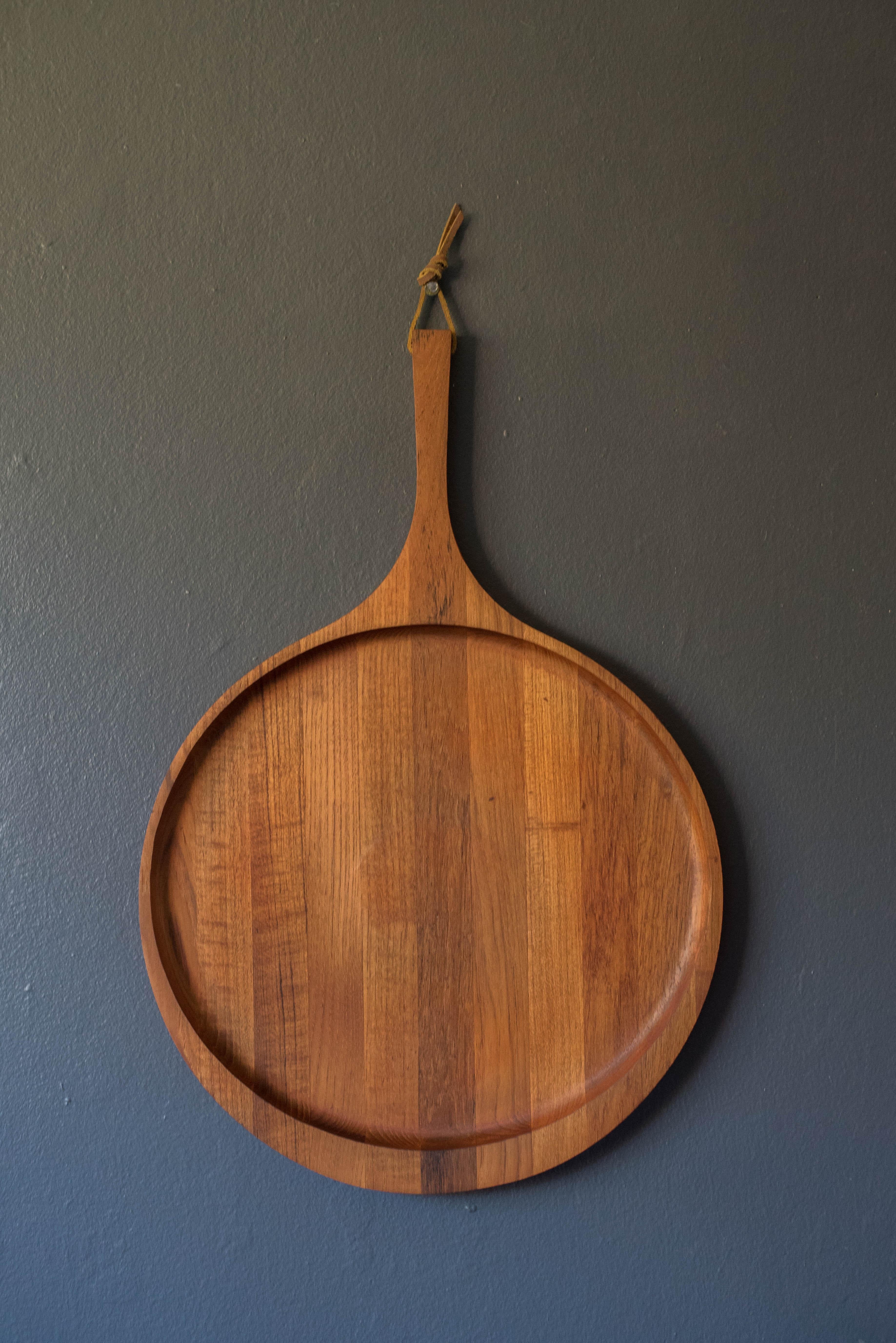 Mid-Century Modern serving platter manufactured by Kjeni, Denmark circa 1960's. This sculptural round tray is crafted in solid planked teak and features a handle with a hanging leather strap perfect to accessorize in the kitchen as a display piece.