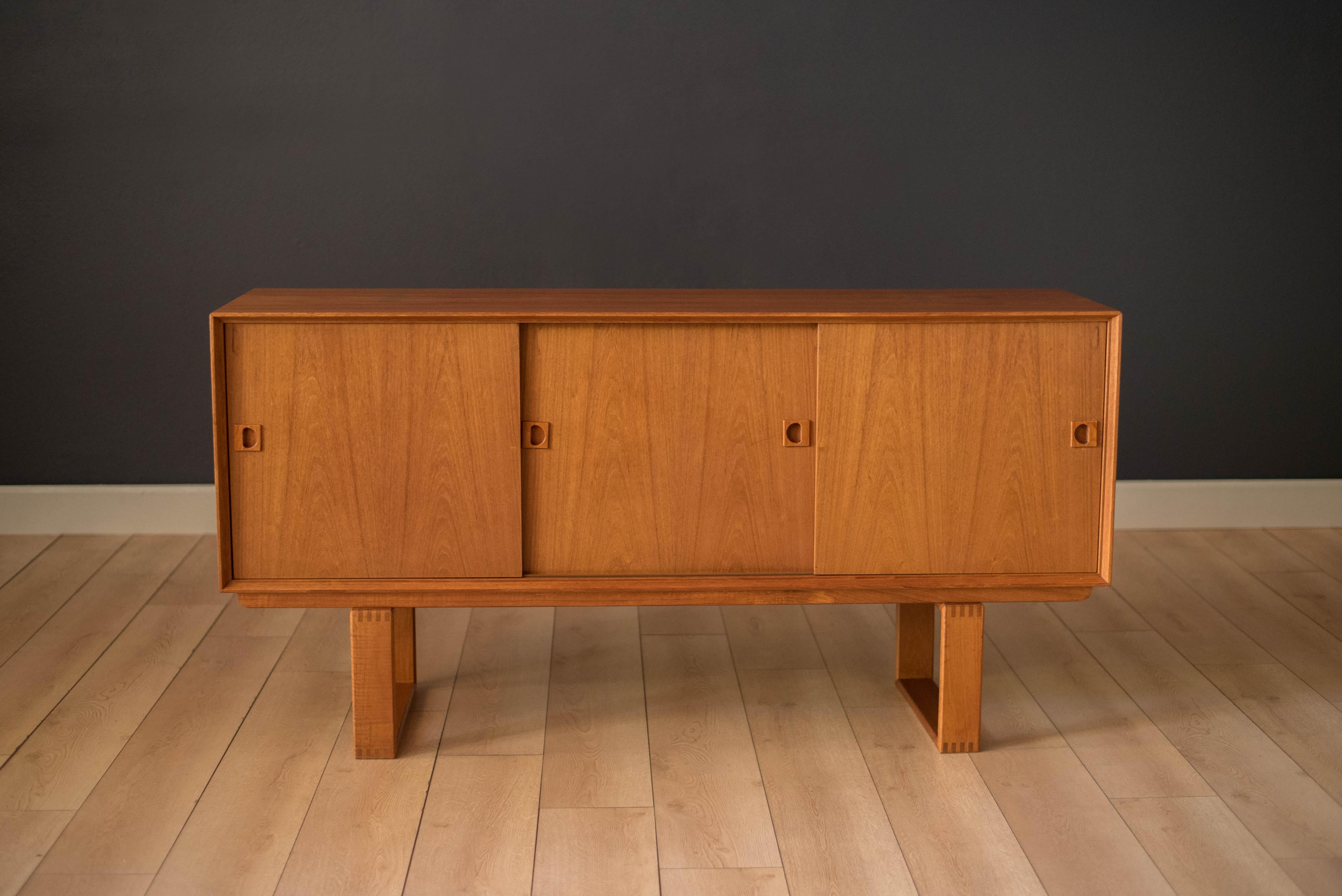Mid-Century Modern sideboard credenza in teak manufactured by Dyrlund, circa 1960s. This piece features sliding cabinet doors with sculpted handles. Includes three dovetailed drawers and open storage compartments with adjustable shelving. Supported