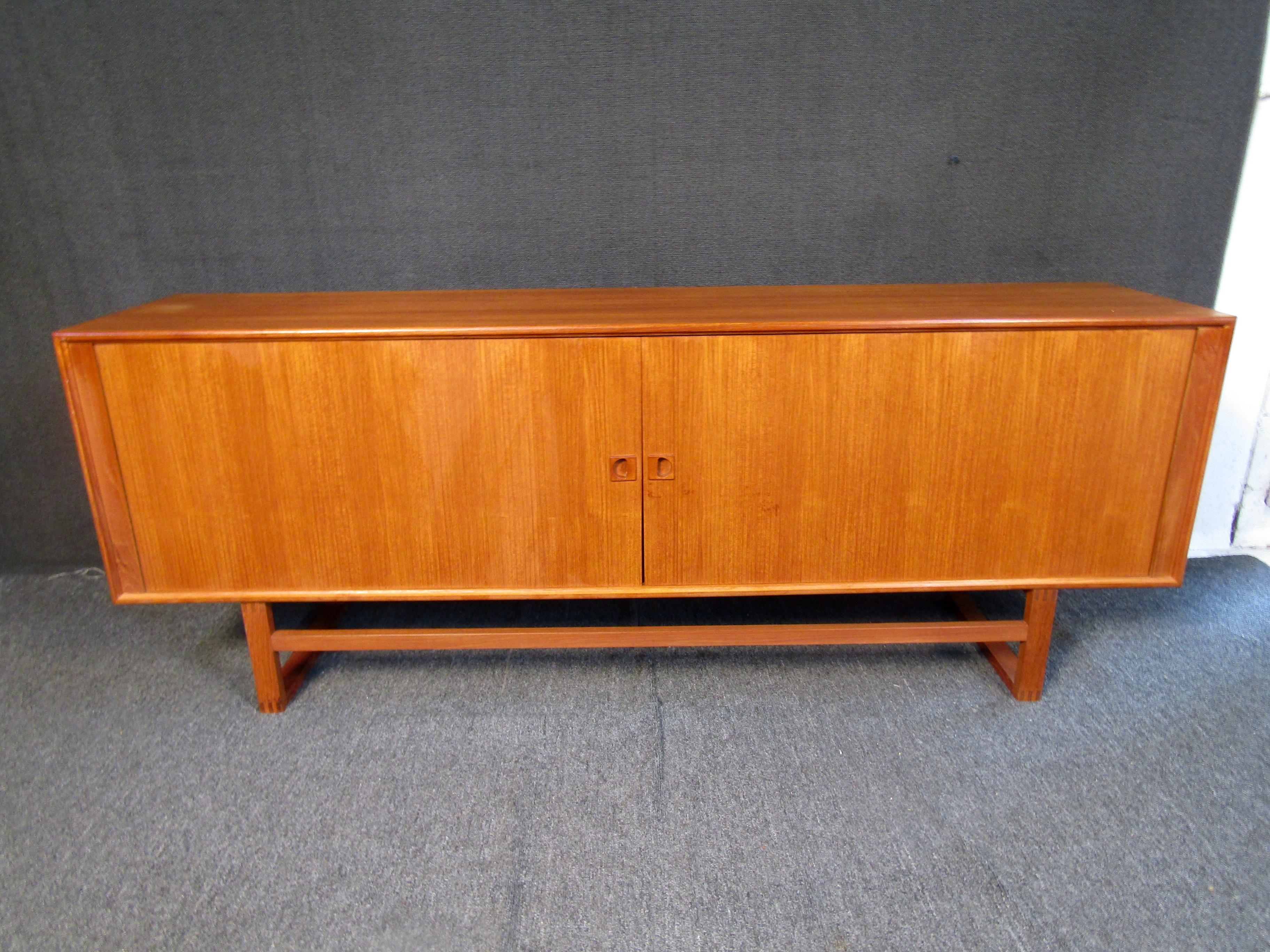 This stunning danish modern tambour door sideboard features a beautiful teak finish, and spacious interior cabinet space. Multiple shelves and a lined center drawer that makes this a versatile storage piece for any Mid-Century setting. Made in