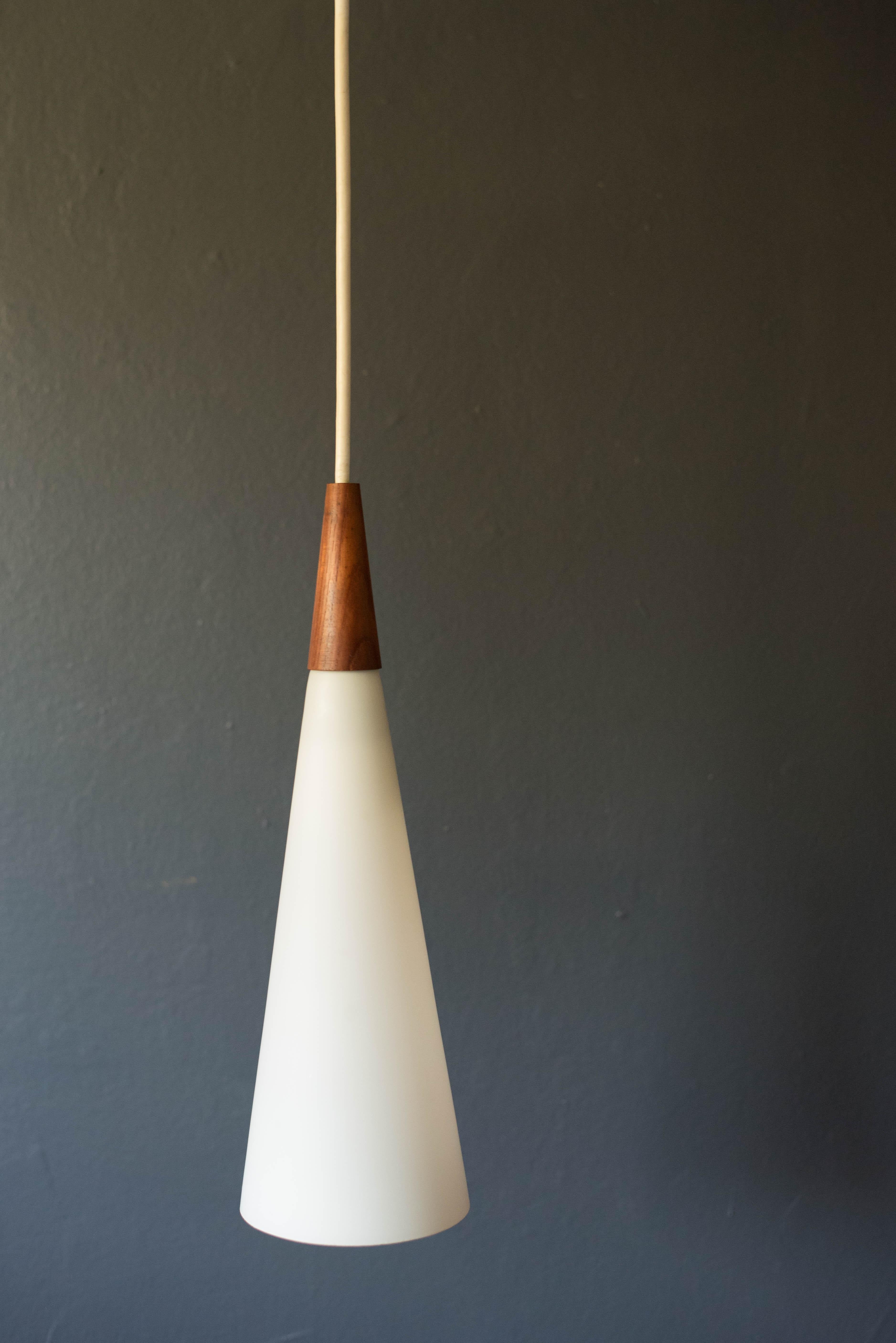 Mid-Century Modern hanging pendant by Holmegaard, circa 1960s. This piece displays a white frosted glass shade that emits a glowing light accented with teak trim.