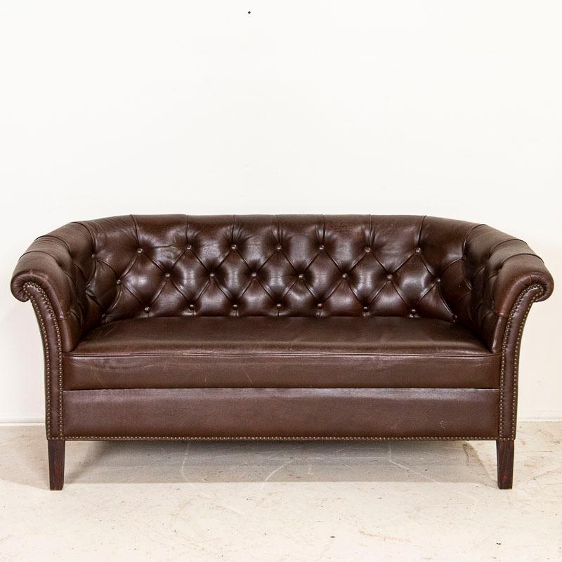 Loaded with character, this small sofa (or loveseat) packs a vintage punch with the classic lines and tufted back of a small scale Chesterfield sofa. The dark brown original leather shows typical age related wear seen in small scuffs/scratches and