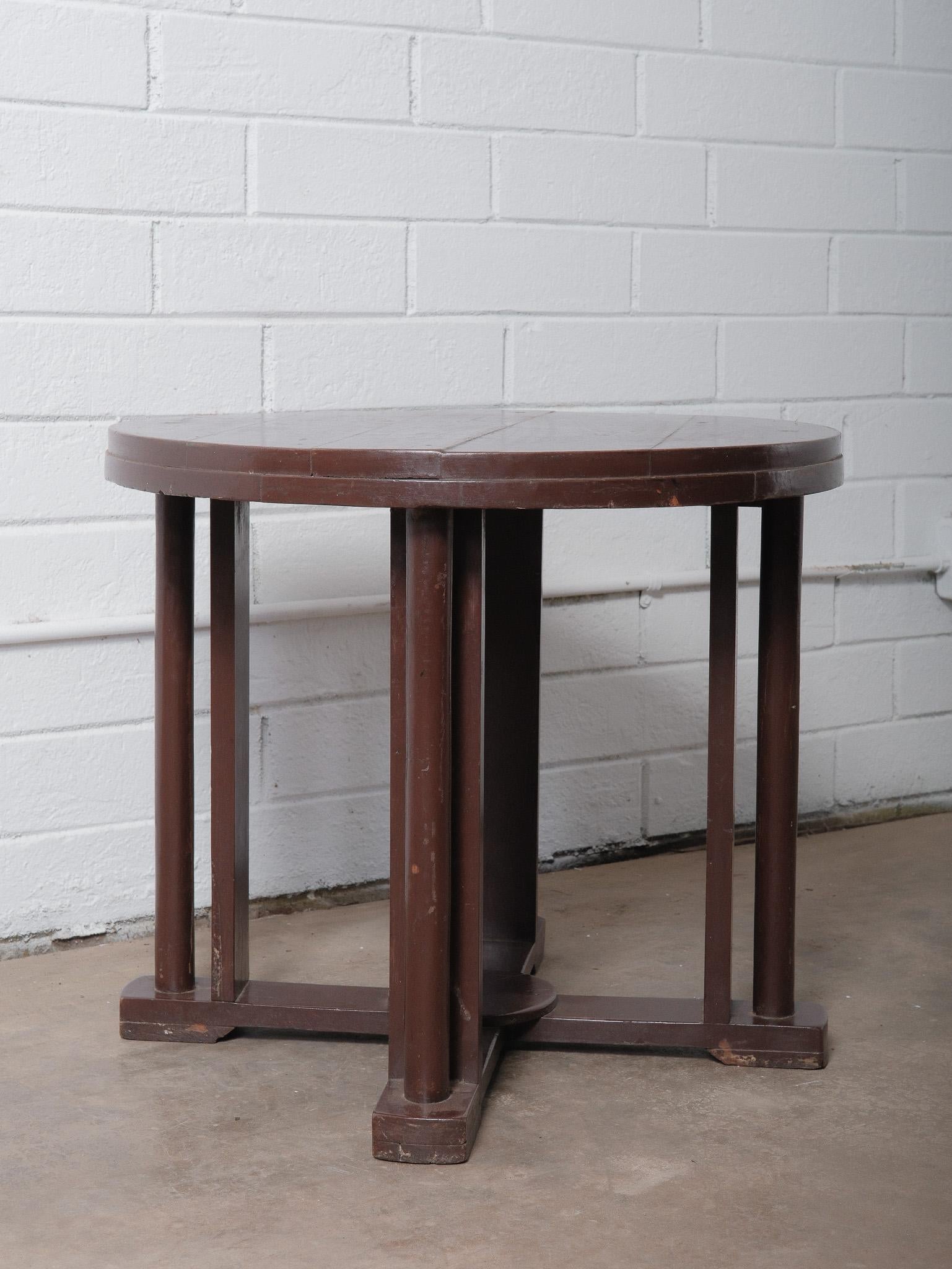 This vintage wooden round side table from India dates to the mid-20th century and features a distinctive architectural base composed of four vertical supports joined by cross-braced feet. The circular top is crafted from solid wood planks, retaining