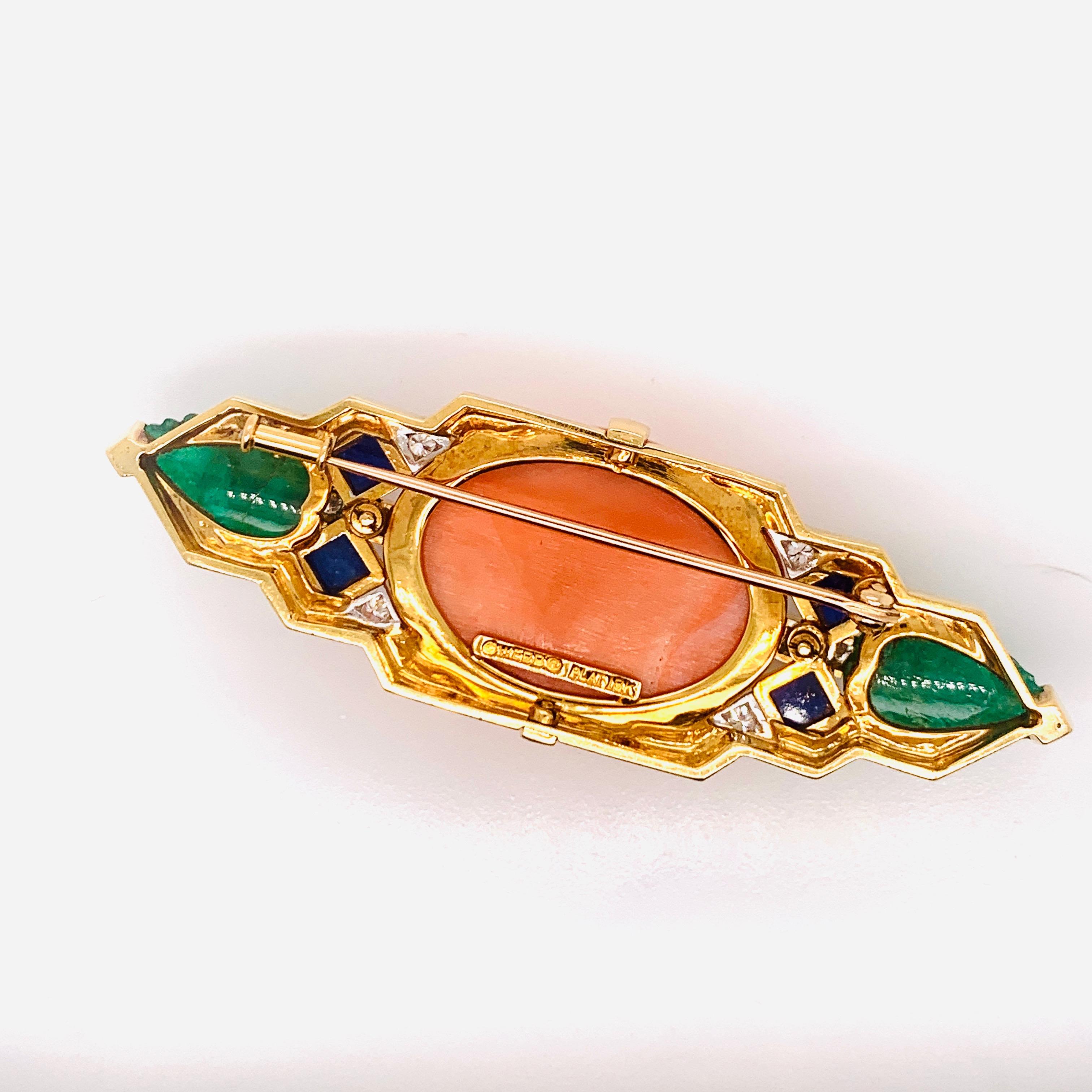 This estate David Webb brooch is the perfect addition to any jewelry collection. The main stone on this brooch is a vibrant oval shaped coral, surrounded by four square cut lapis lazuli, ten round cut diamonds, and two emeralds. All of these