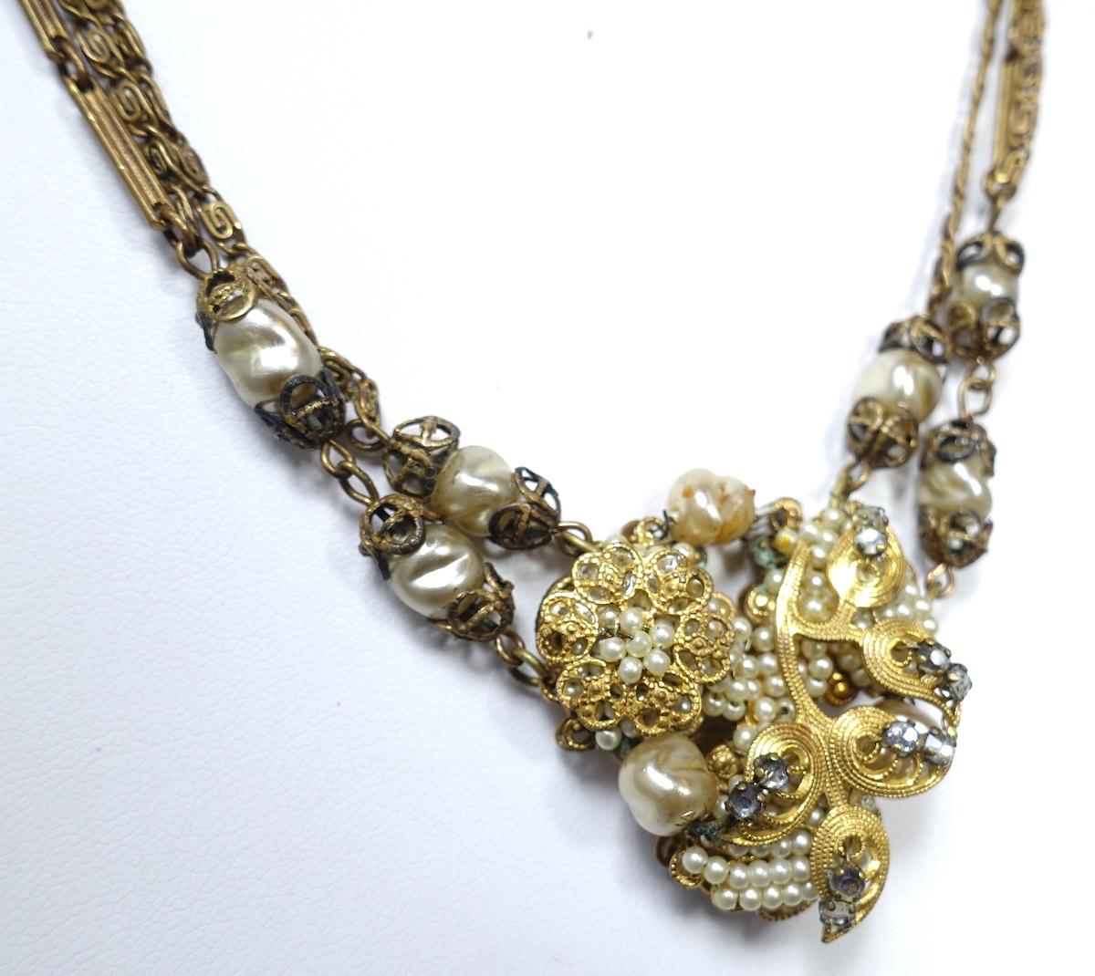This vintage early Miriam Haskell necklace features faux pearls with rose montee accents in a gold tone setting. It has a double chain with a slide in clasp.  The necklace measures 16-1/2” with the centerpiece 1-1/2” x 1-3/8”. This necklace is in