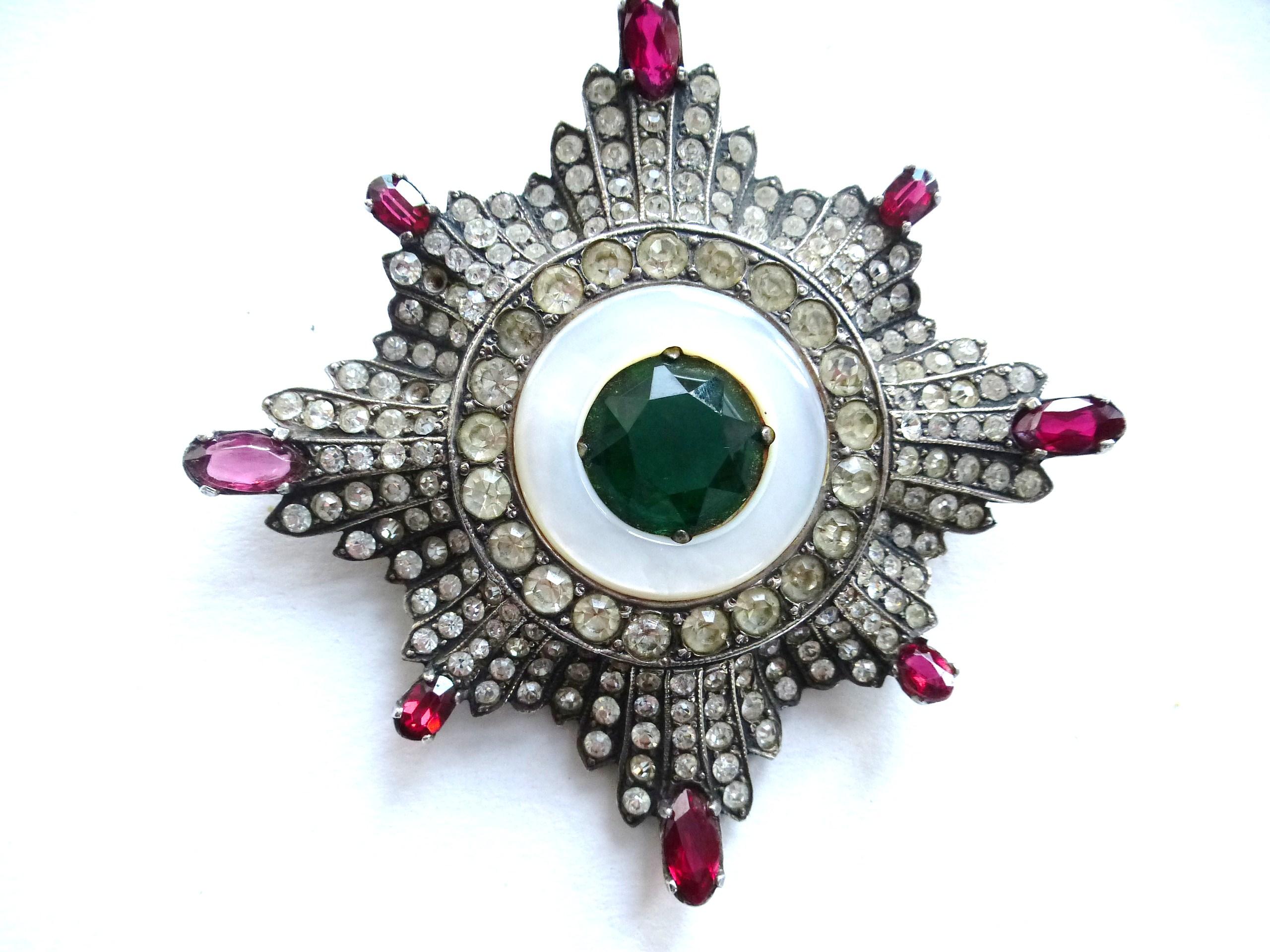 A very early star brooch completely filled with many different large cut rhinestones. The stones get bigger towards the inside, then a mother-of-pearl ring surrounds a large, cut green rhinestone. the outer tips are adorned with red rectangular