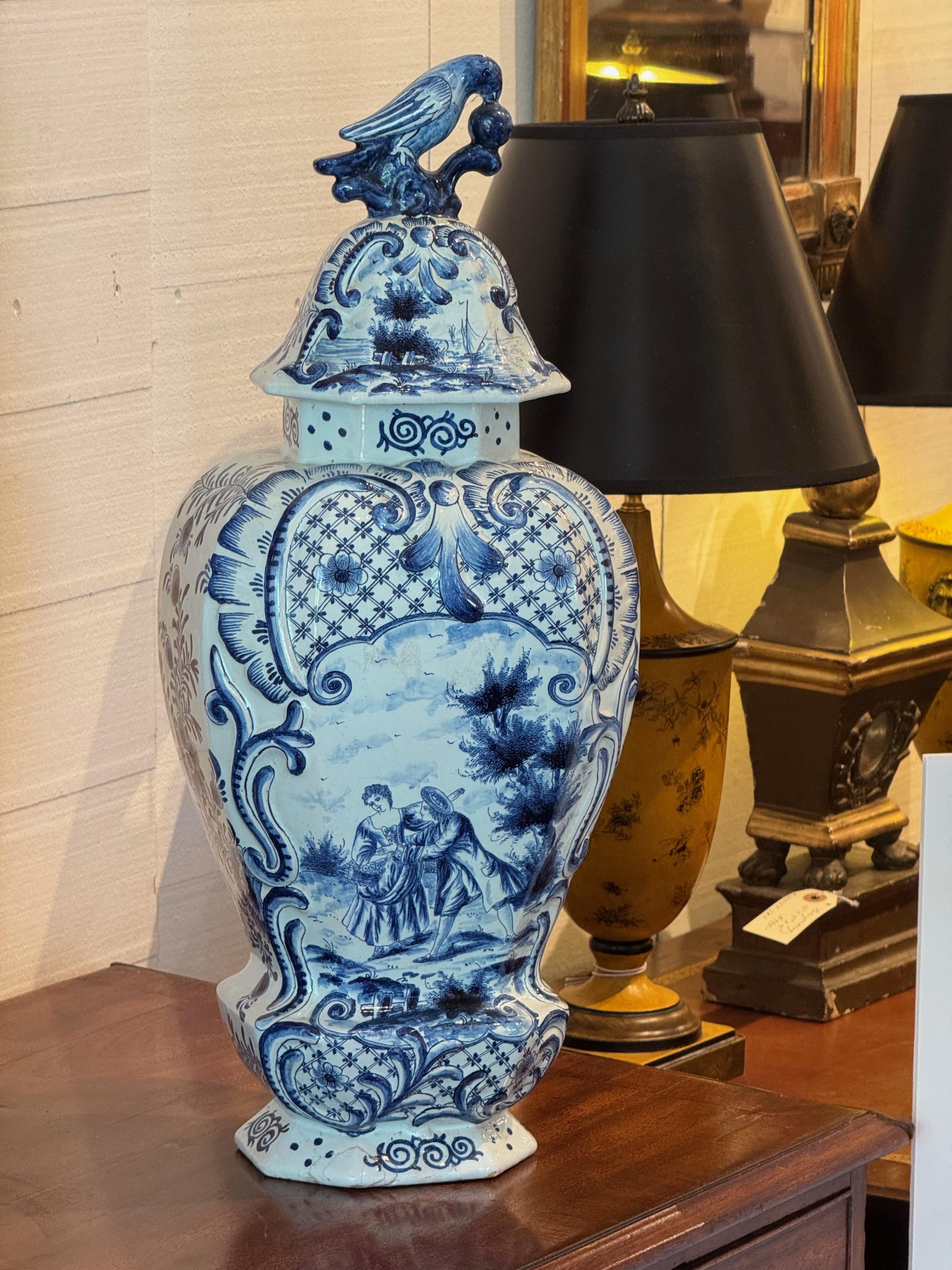 Blue and white hand-painted Delft urn with a classic figural pastoral scene on the body and ornate scrollwork throughout. The lid features a high-relief bird finial holding fruit in its beak
