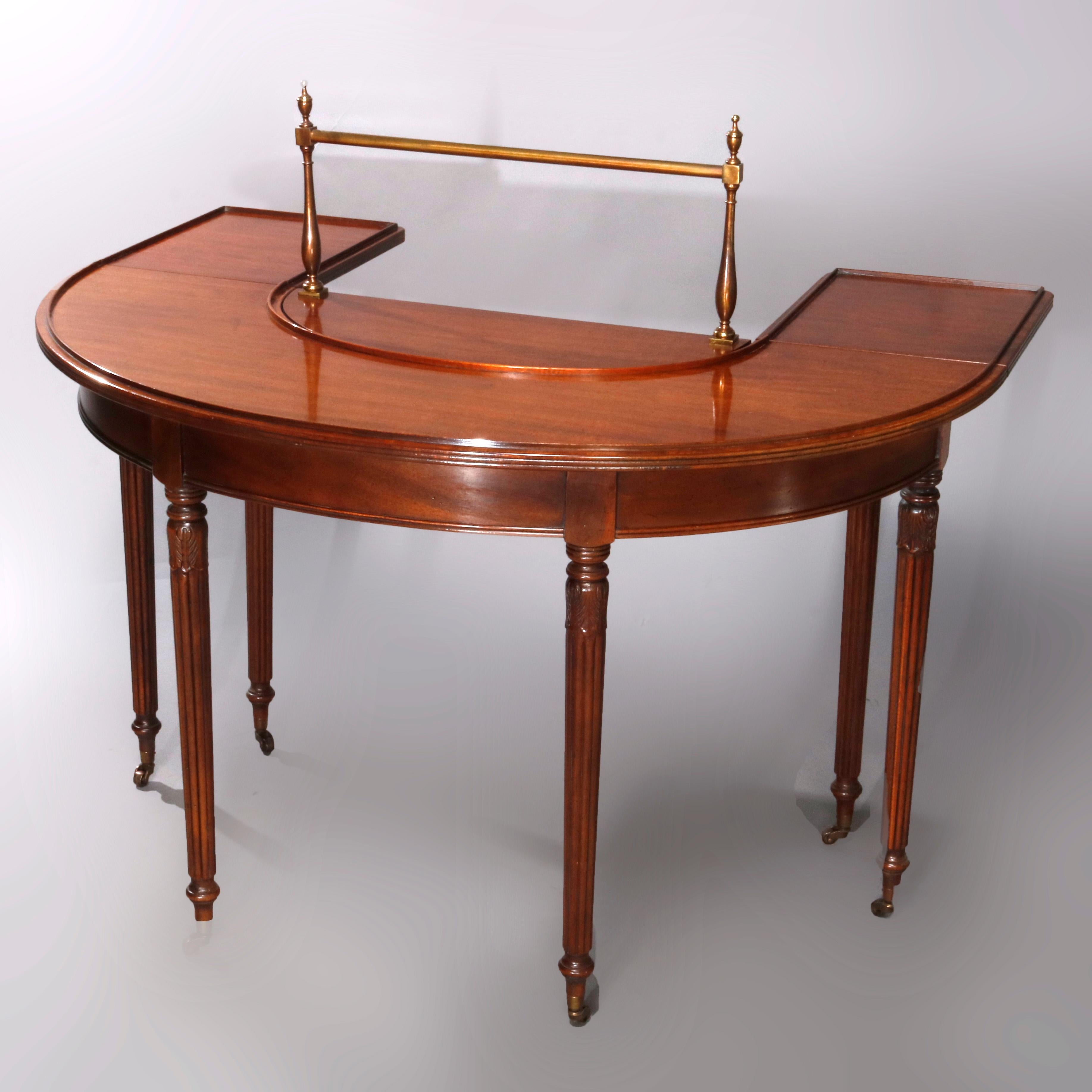 A vintage Sheraton style serving table by Drexel Furniture Co. of the Wallace Nutting collection offers mahogany construction in wraparound demilune form with rear drop leaves and brass linen rail, raised on reeded and tapered legs having foliate