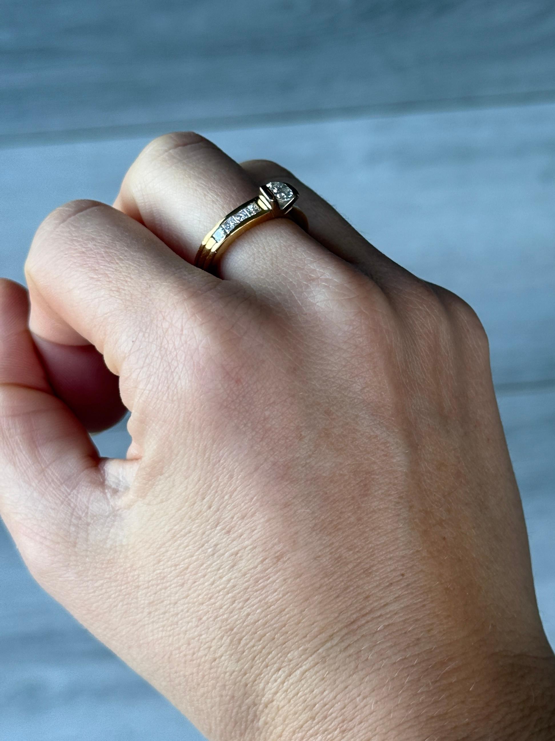 This simply stunning ring holds a clean and bright diamond measuring approximately 35pts. The Diamond is set in a simple claw setting and  sits proud when worn. The ring is modelled out of 18ct gold and have four princess cut diamonds per side.