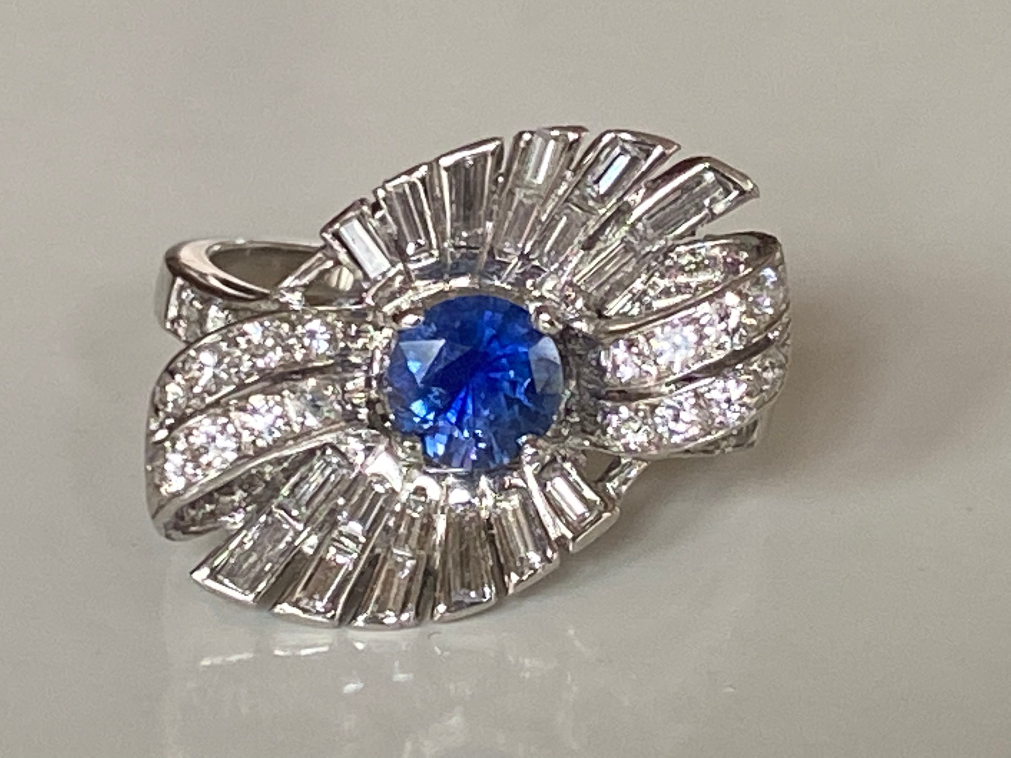 This bold vintage cocktail ring features two ribbons of round-brilliant diamonds and baguette diamonds fanned out around a natural royal blue round sapphire center stone handcrafted in platinum. The diamonds total approximately 2.00 carats. 


