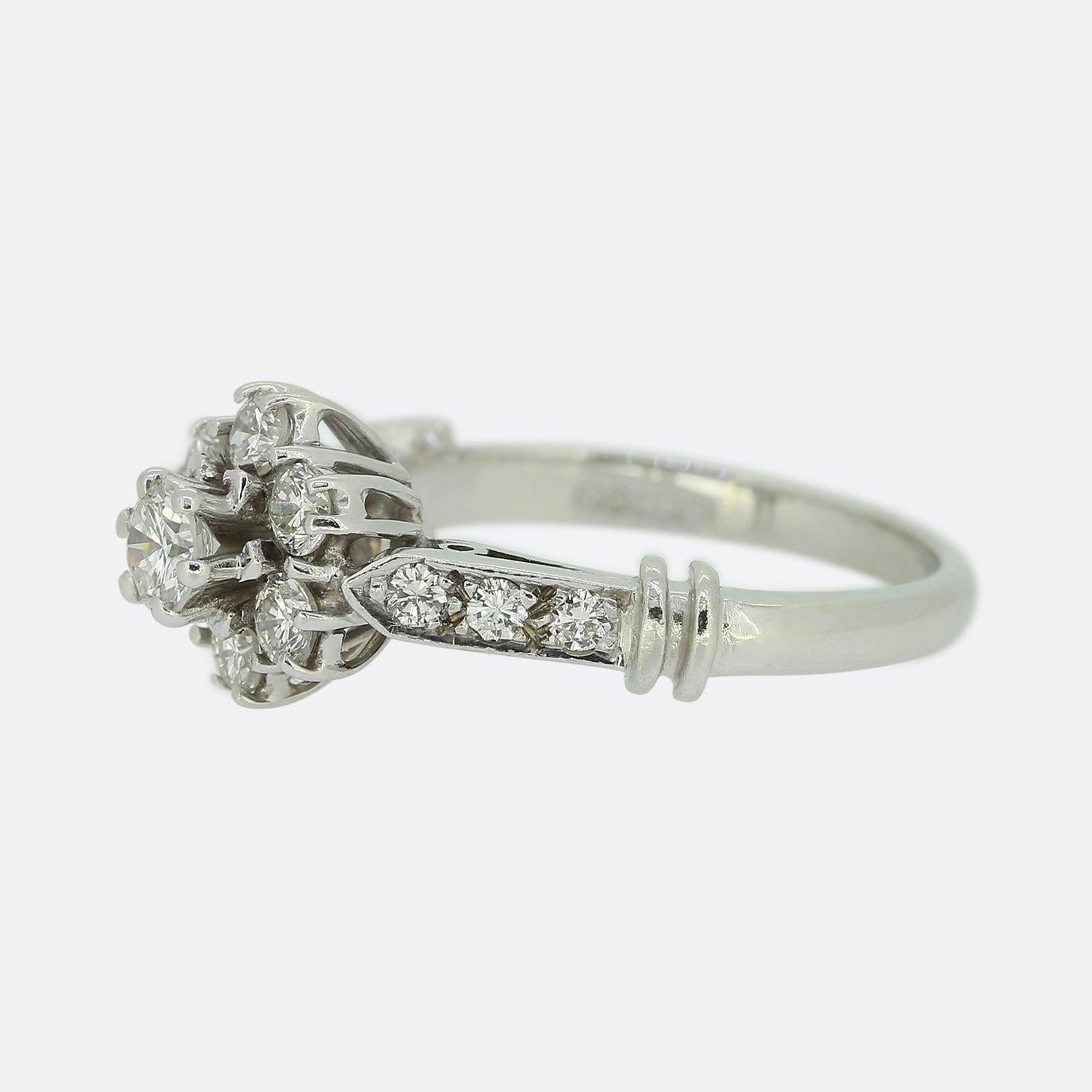 Hier haben wir einen wunderschönen Diamant-Cluster-Ring. Dieses Vintage-Stück wurde aus 18 Karat Weiß gefertigt und zeigt einen einzelnen, leicht erhöhten runden Diamanten im Brillantschliff in der Mitte der Vorderseite. Dieser Hauptstein wird dann