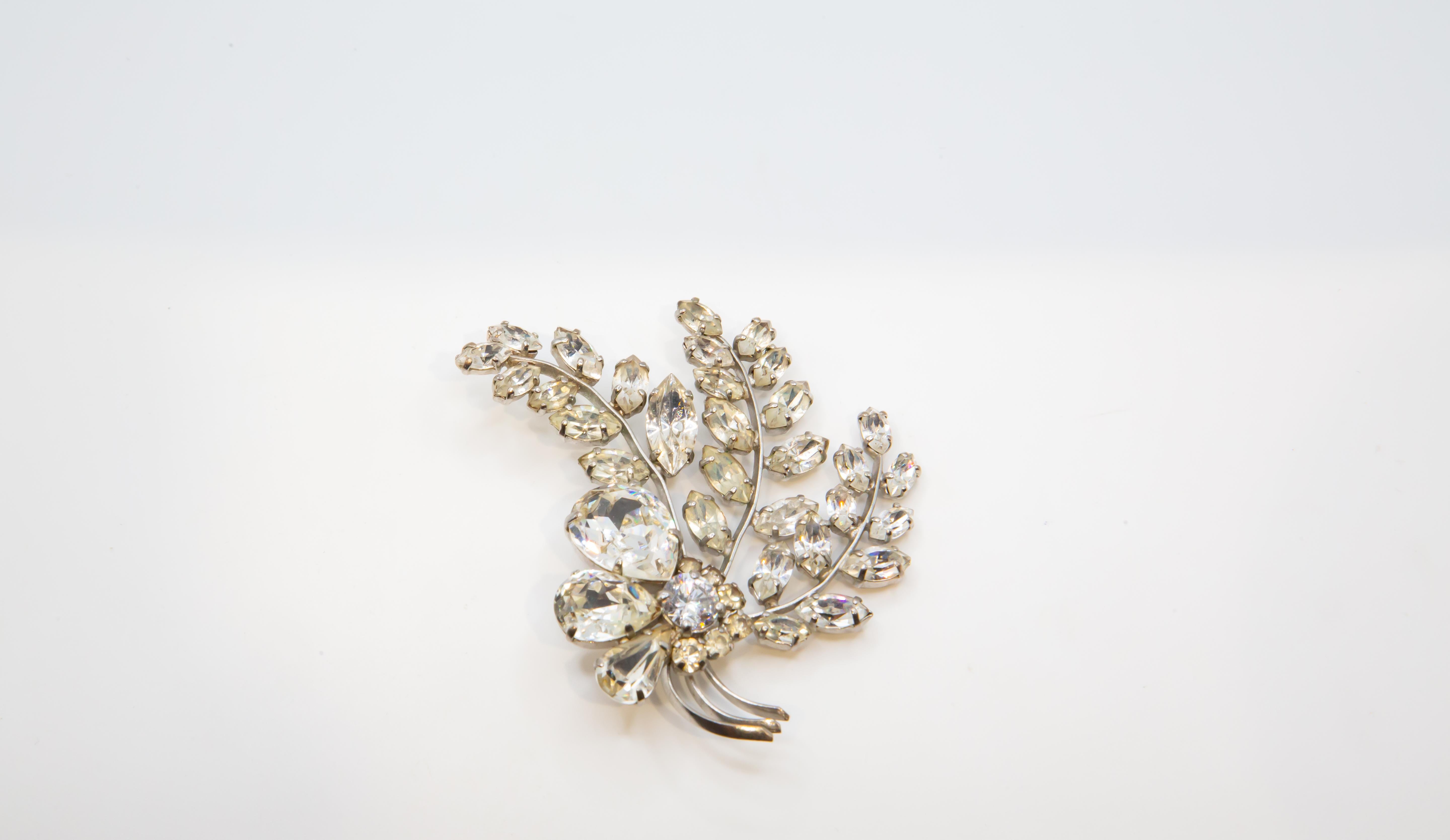 Un'affascinante spilla vintage a forma di spruzzo floreale o bouquet di foglie, impreziosita da strass trasparenti a taglio brillante o diamanti simulati incastonati in metallo argentato. Ogni 