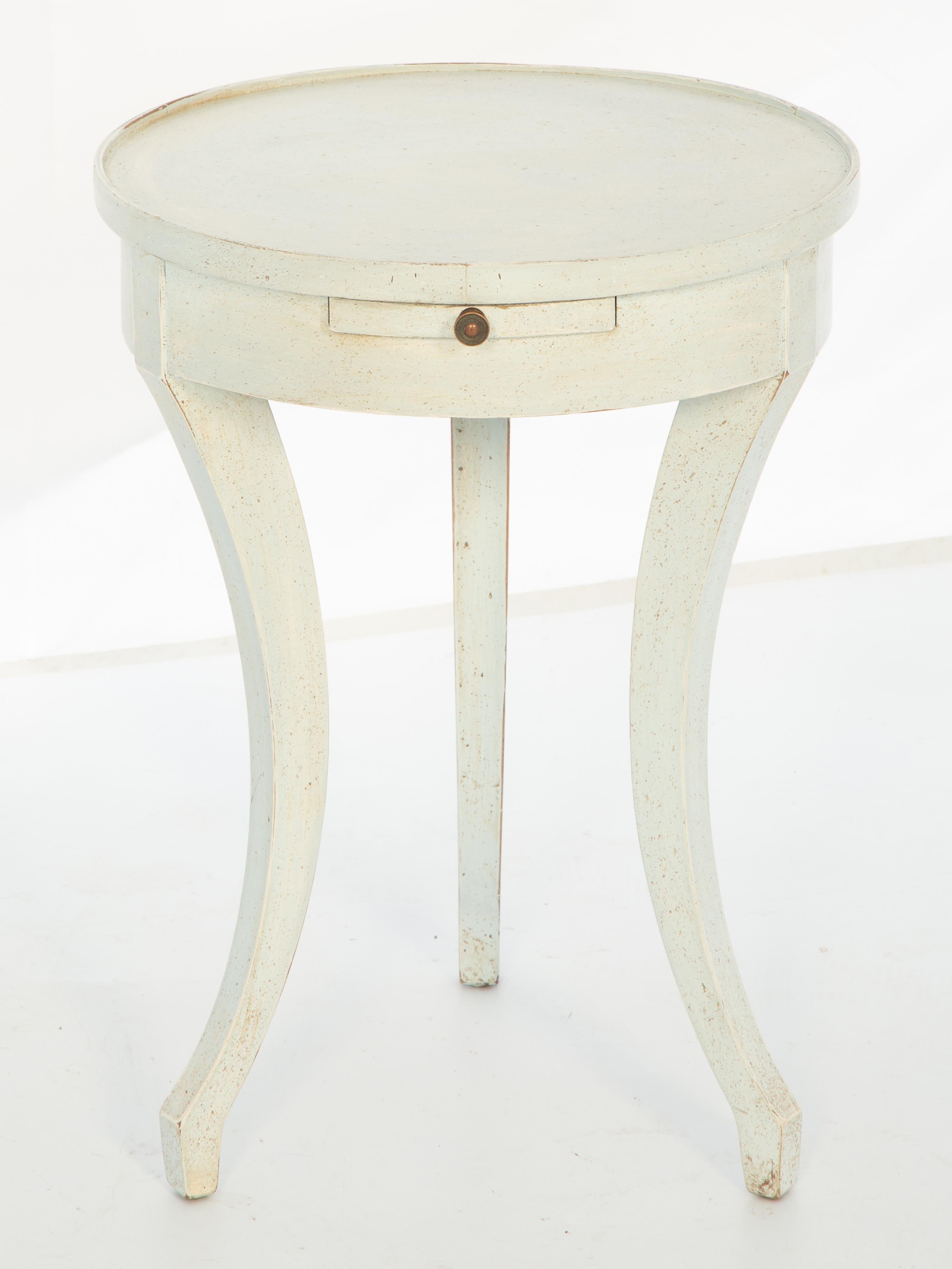 Vintage Directoire-style accent table, by Drexel, having an ivory painted finish showing natural wear. Its round top over an apron containing a slide, raised on a trio of sabre legs.

Stock ID: D4037