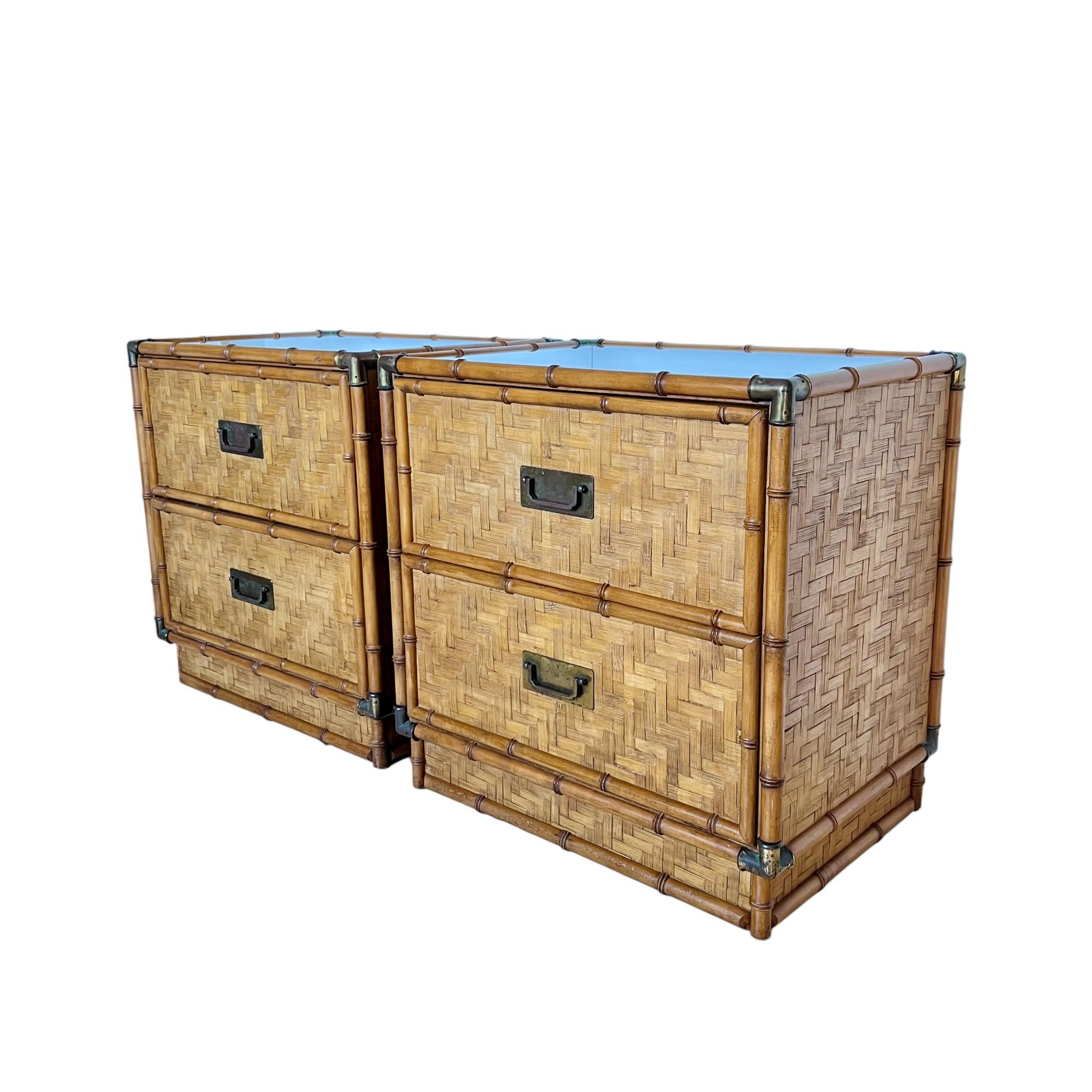 A vintage pair of mid-century modern campaign style two-drawer nightstands or commodes produced by Dixie Furniture, circa 1965. They feature woven split reed paneling with turned wood faux bamboo framing, and are accented with brass handles and