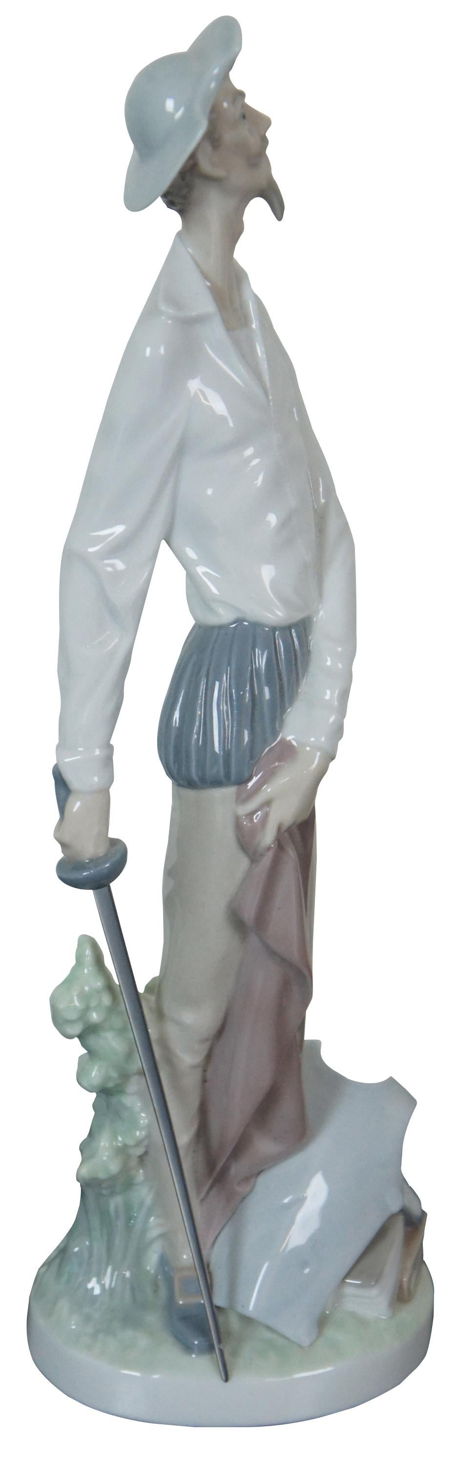 Retired Lladro Spain porcelain figurine of Miguel de Cervantes’ Don Quixote standing with a silver rapier, holding a red cape with chest plate and several books at his feet. #4854.
  