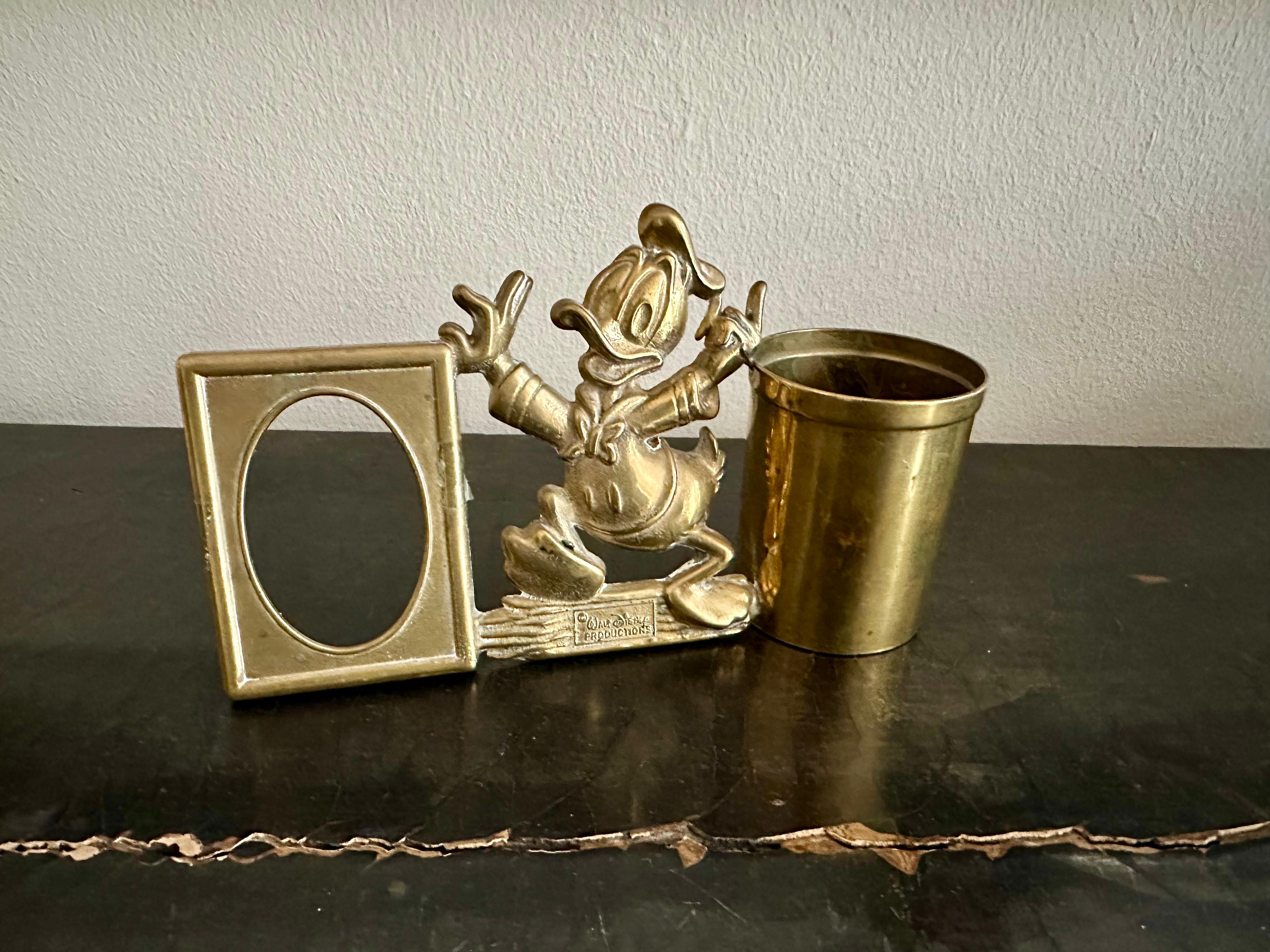 This charming vintage pen holder with a built-in picture frame features the beloved Donald Duck, one of the most iconic Disney characters. Dating back to the 1970s, this collectible piece is crafted from solid brass, giving it a timeless and durable