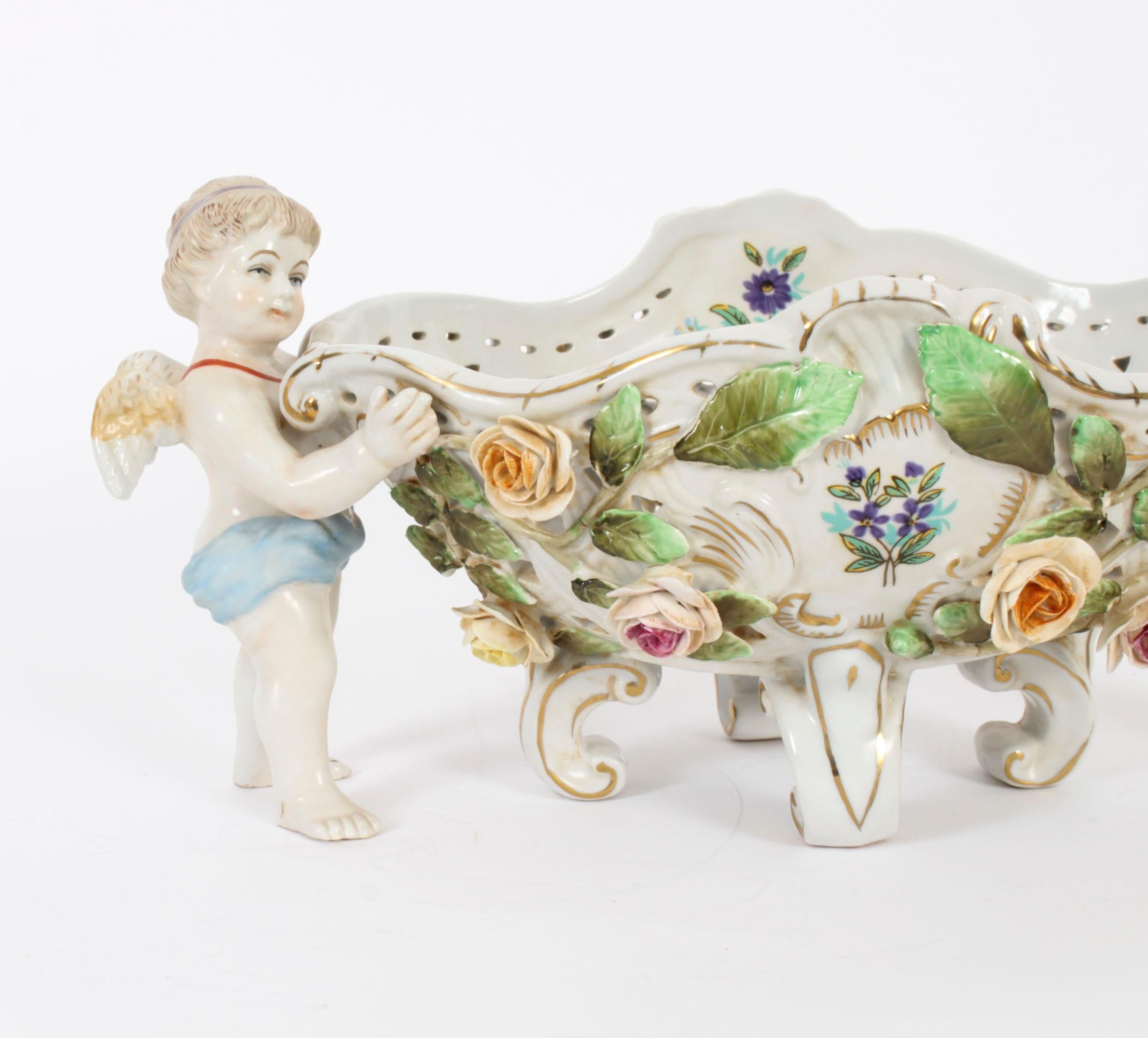 This is a beautiful and ornate Vintage Dresden Revival porcelain centrepiece, late 20th century in date.

The centrepiece features a winged cherub on each side  holding an oval pierced basket-like dish with hand painted roses and foliate decoration