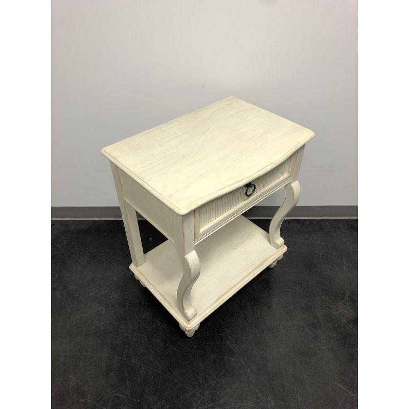 A Cottage style nightstand by Drexel Heritage, their Chelsea Cottage. Solid hardwood painted an off-white color with brass hardware. Features one drawer of dovetail construction and an undertier open shelf. Made in North Carolina, USA, in the late