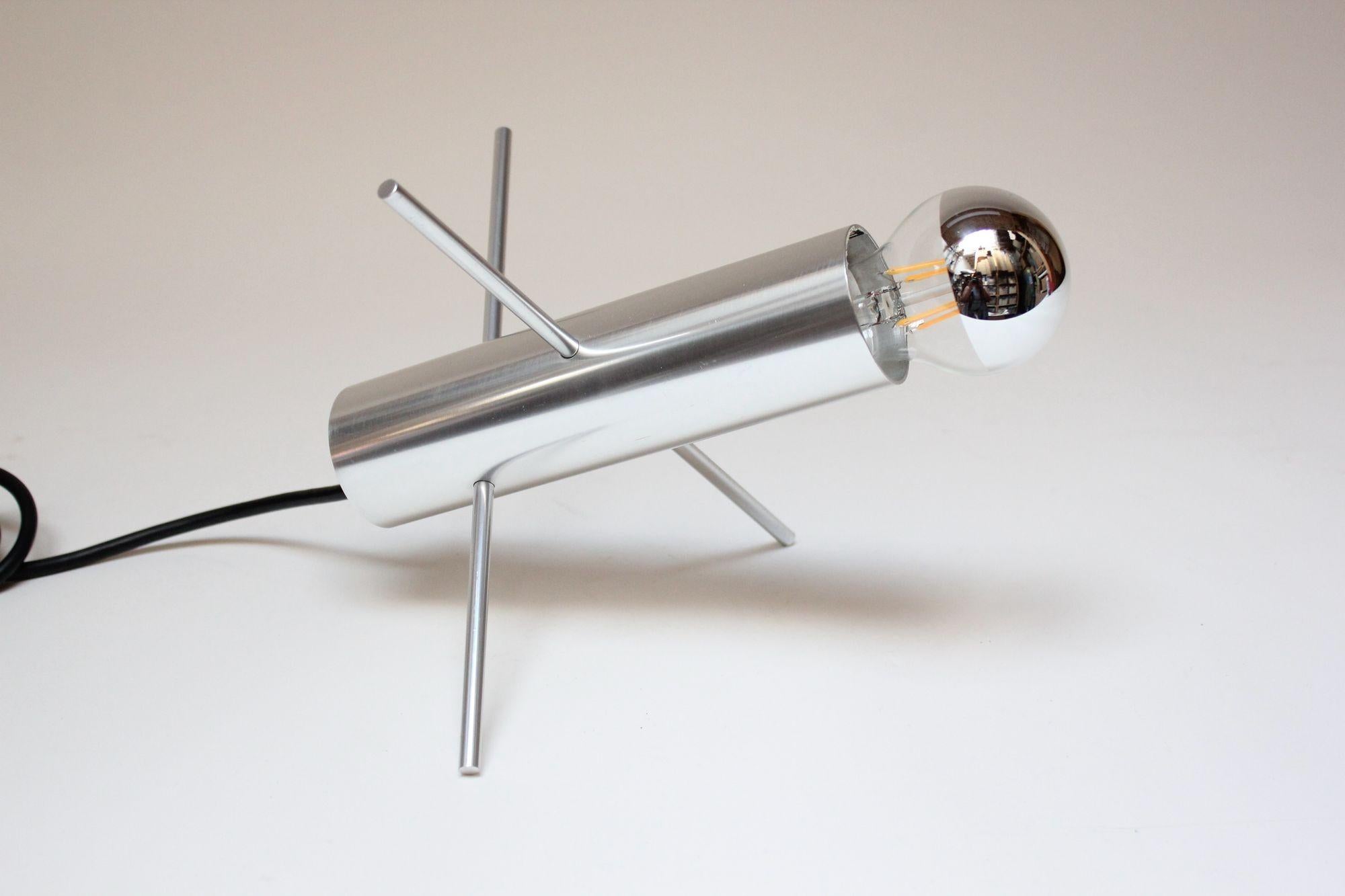 Lampe de table 'Krekel' / 'Cricket' de forme modeste, modèle R-60 conçu par Otto Wach pour RAAK (Hollande, 1962).
Composé d'un cylindre en aluminium brossé avec quatre perforations abritant deux tiges/épingles en aluminium entrecroisées qui