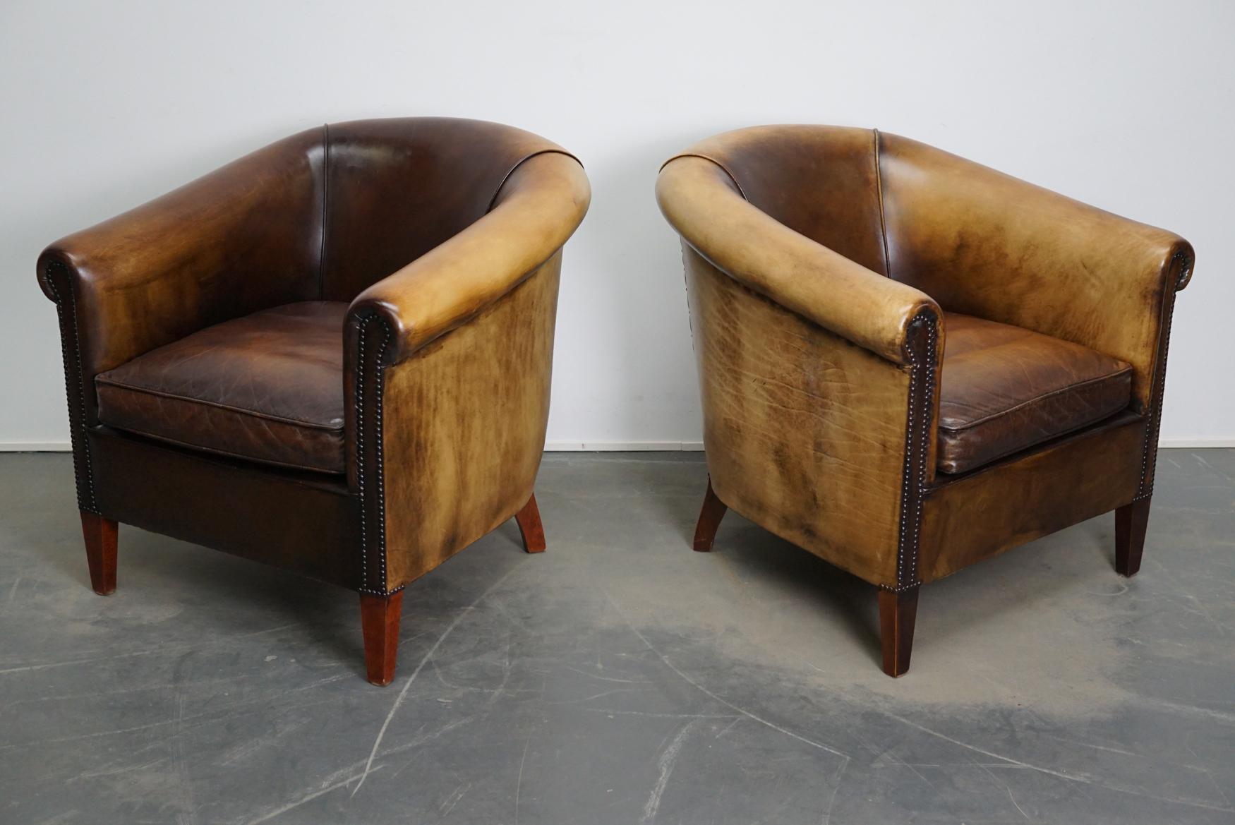 This pair of brown-colored leather club chairs come from the Netherlands. They are upholstered with cognac-colored leather and feature metal rivets and wooden legs.