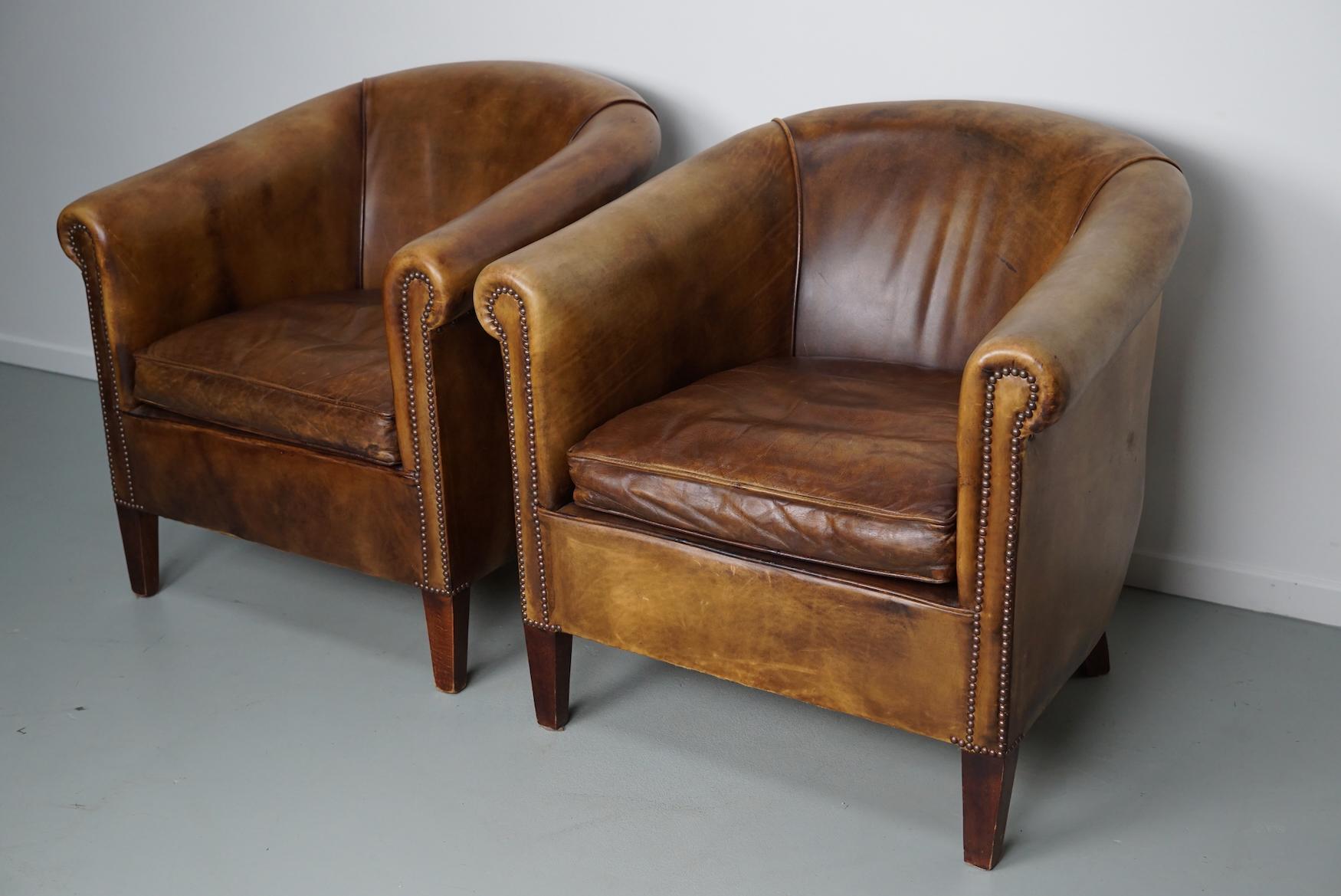 This pair of cognac-colored leather club chairs come from the Netherlands. They are upholstered with cognac/brown-colored leather and feature metal rivets and wooden legs.