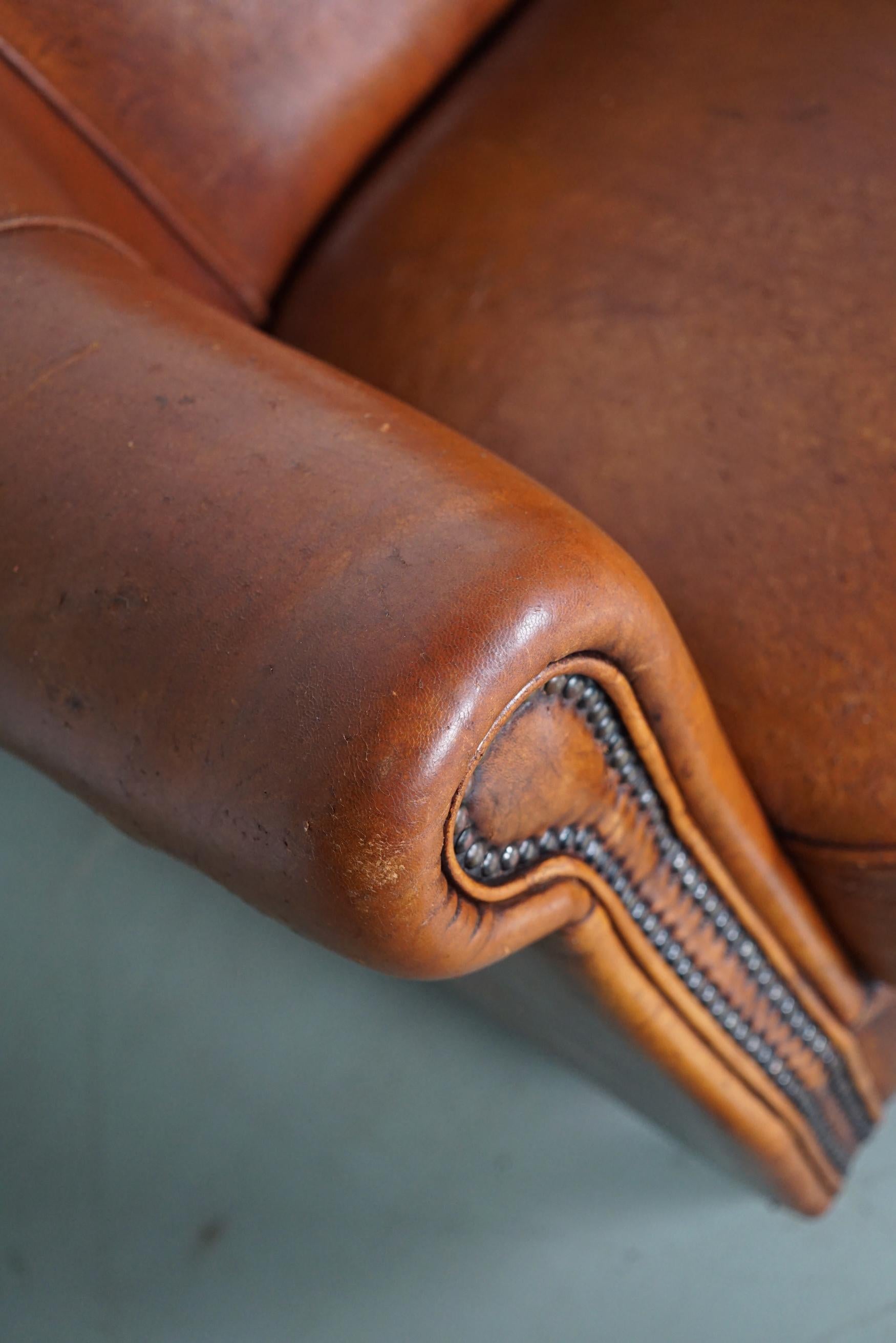 Este sillón club de cuero color coñac procede de Holanda. Está tapizado en piel de color coñac y tiene remaches metálicos y patas de madera.