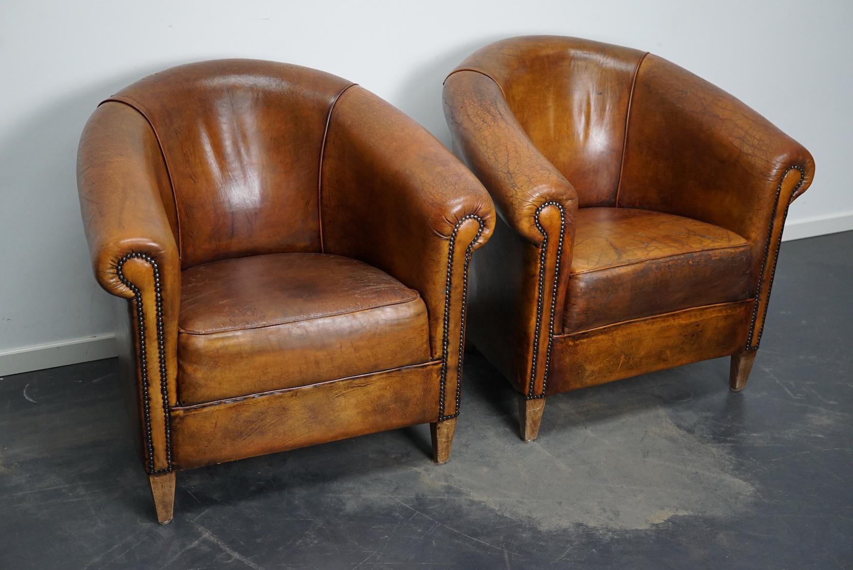 This pair of cognac-colored leather club chairs come from the Netherlands. They are upholstered with cognac-colored leather and feature metal rivets and wooden legs. The chairs show some damage / cracking of the top layer.