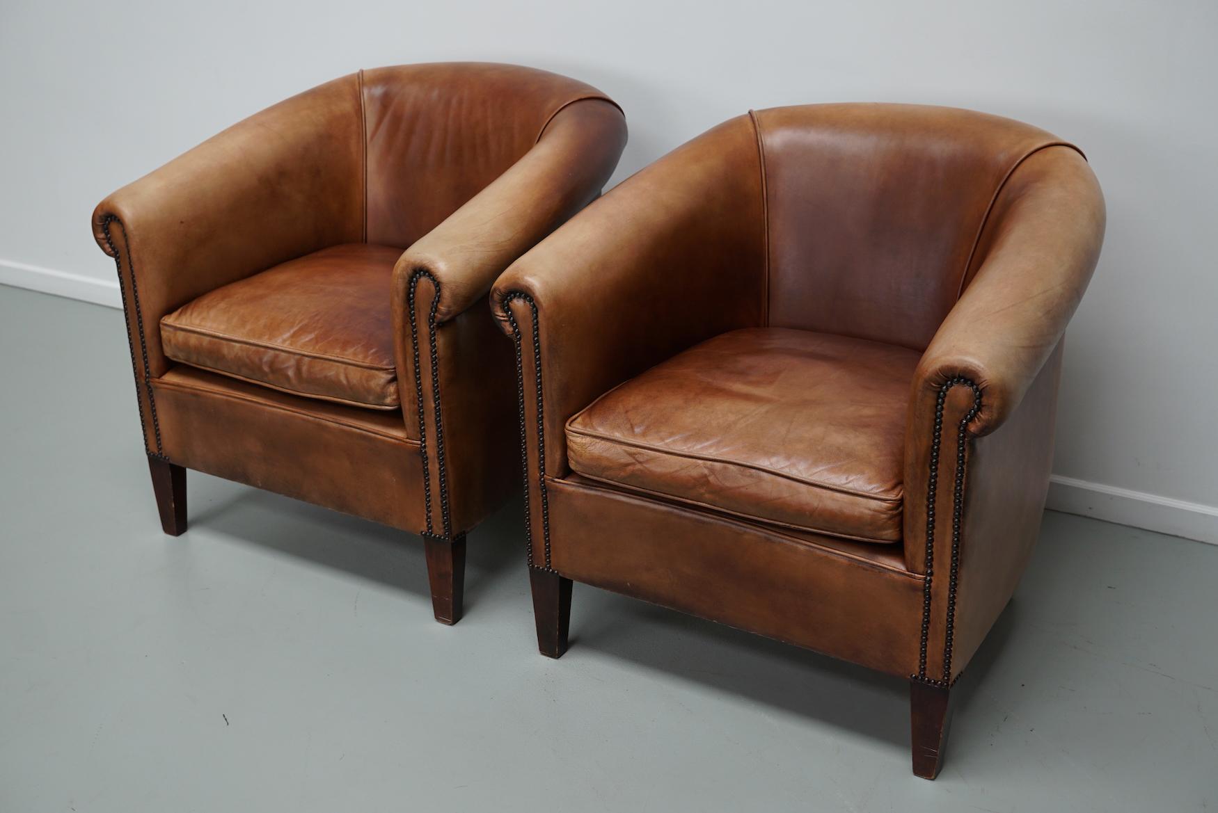 This pair of cognac-colored leather club chairs come from the Netherlands. They are upholstered with cognac-colored leather and feature metal rivets and wooden legs.