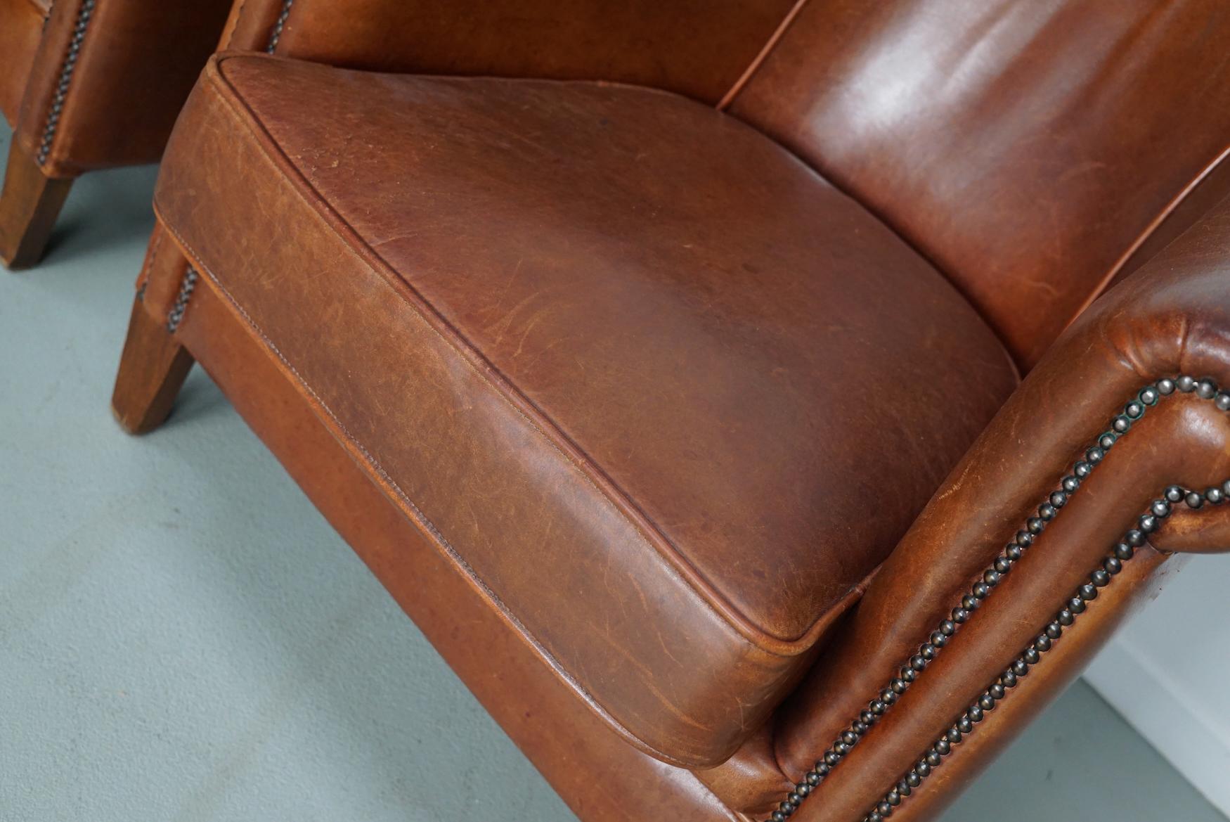 This pair of cognac-colored leather club chairs come from the Netherlands. They are upholstered with cognac-colored leather and feature metal rivets and wooden legs.