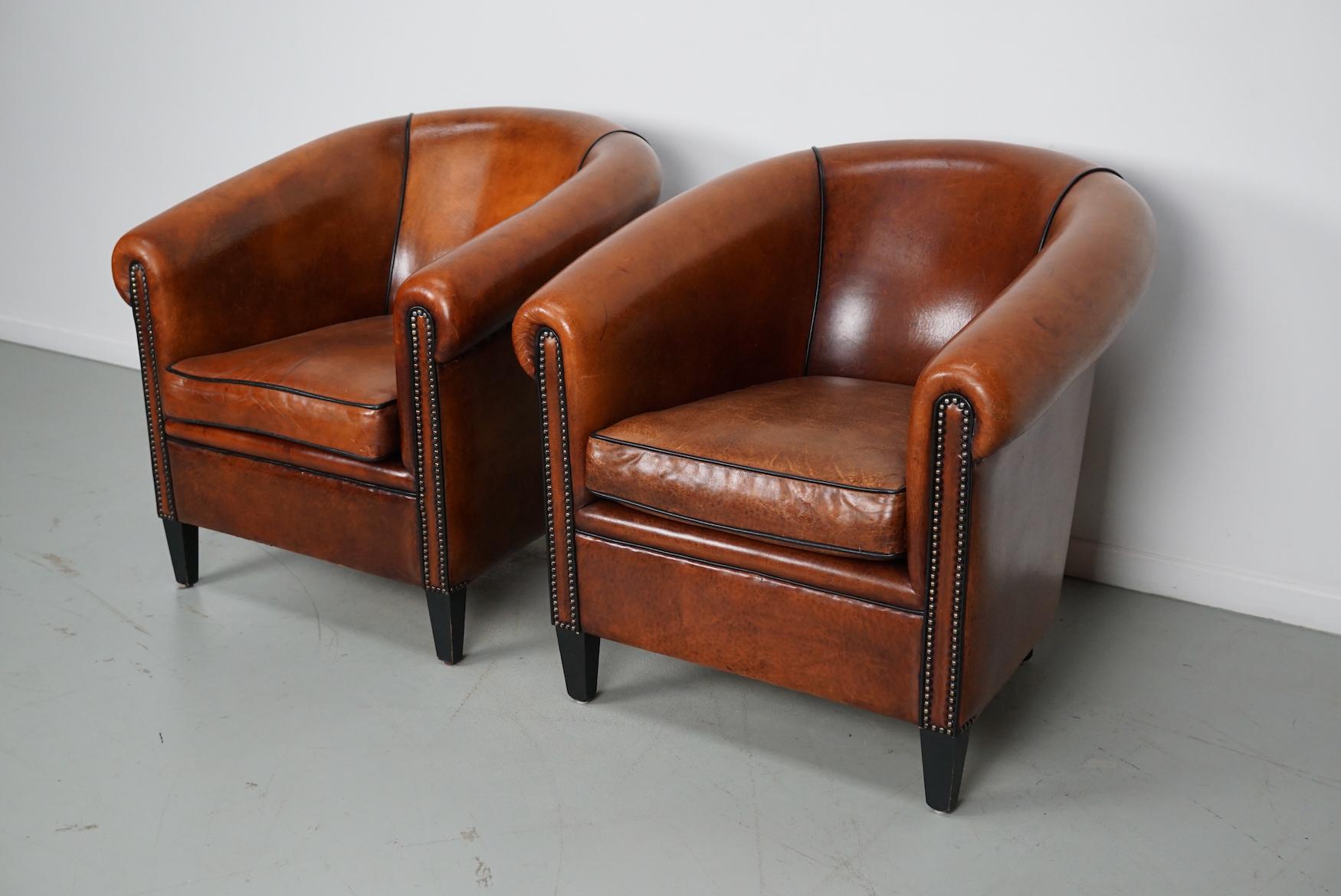 This pair of cognac-colored leather club chairs come from the Netherlands. They are upholstered with cognac-colored leather and feature metal rivets and wooden legs.