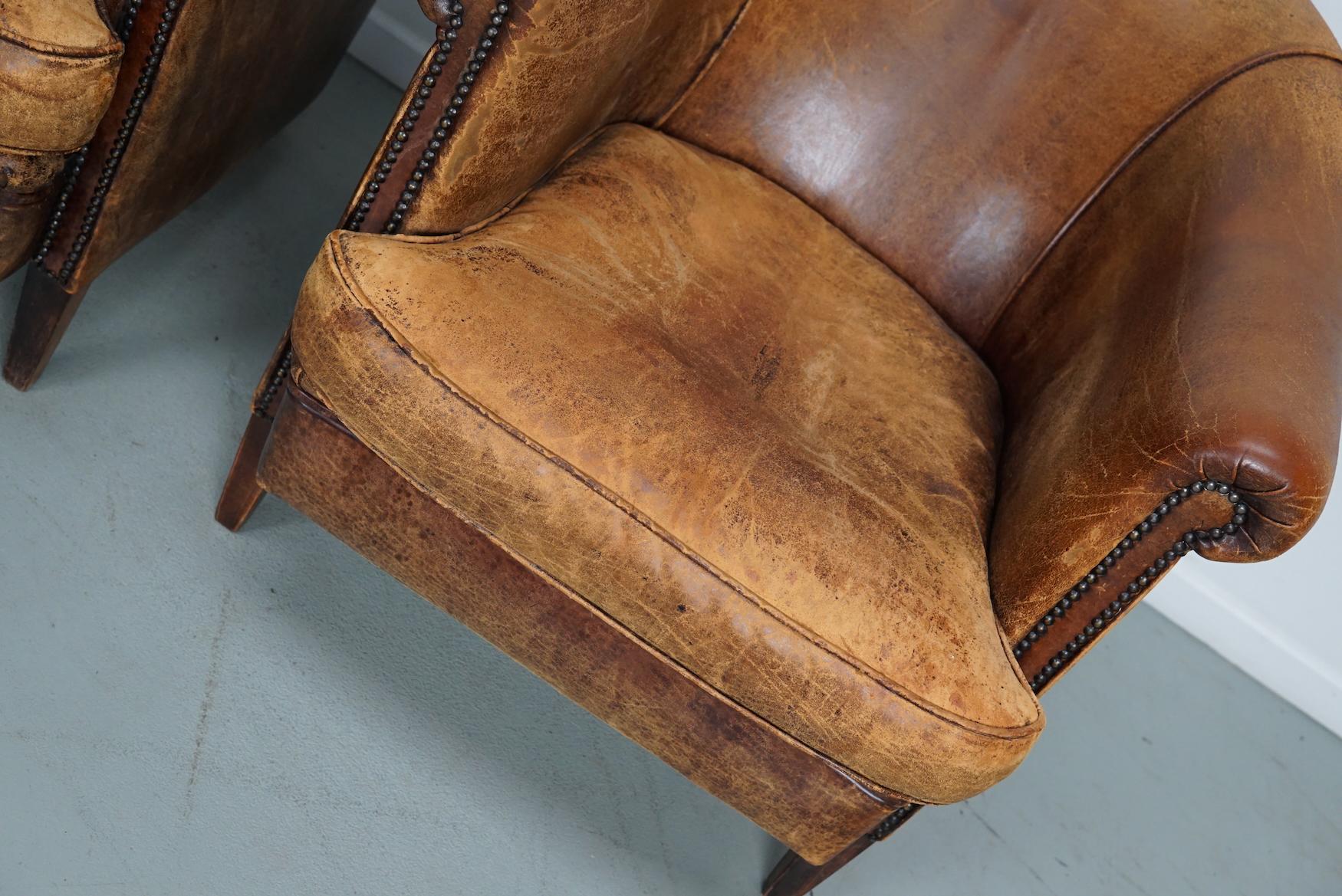 This pair of cognac-colored leather club chairs come from the Netherlands. They are upholstered with cognac-colored leather and feature metal rivets and wooden legs.