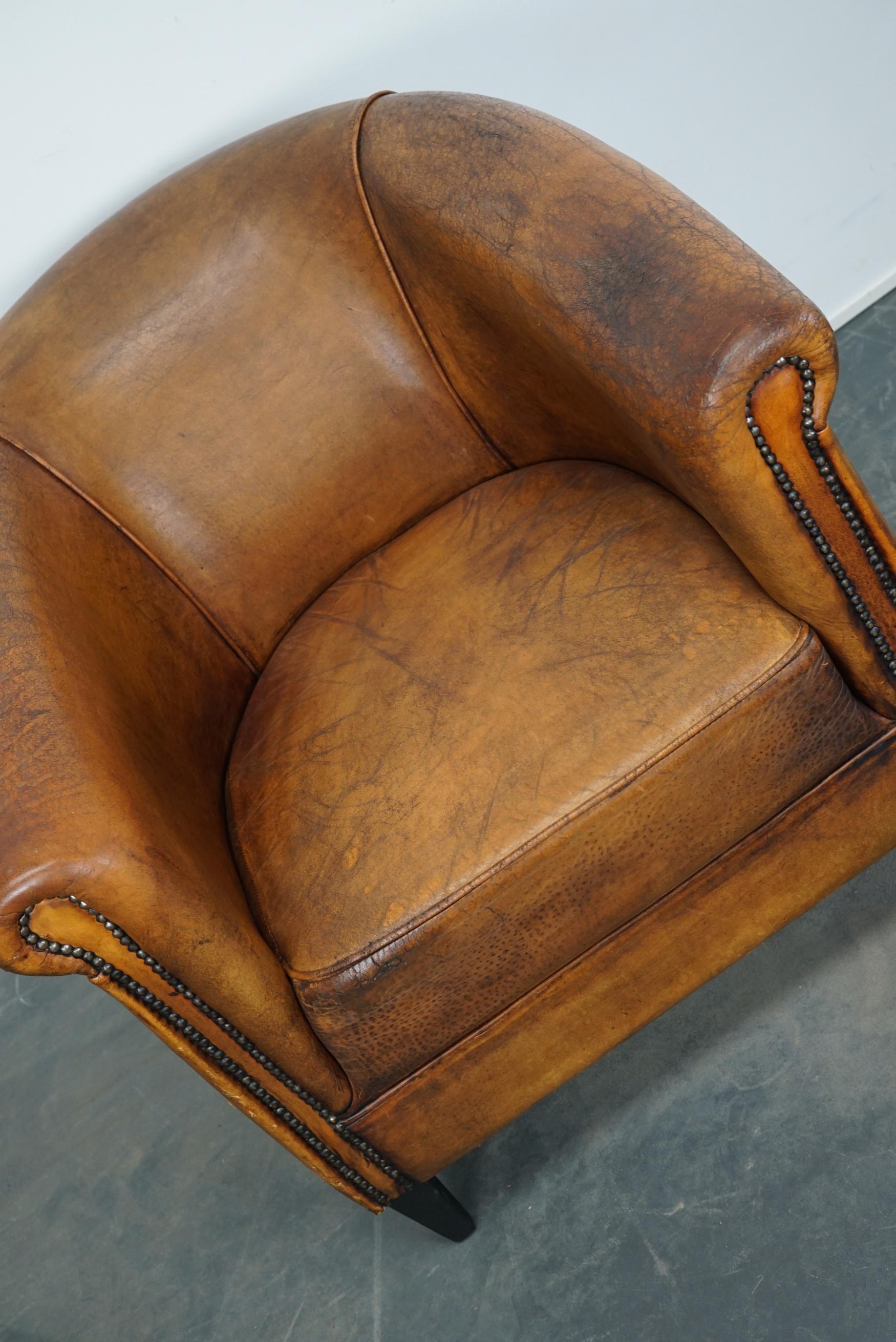 This vintage cognac-colored leather club chair comes from the Netherlands. It is upholstered with cognac-colored leather and features metal rivets and wooden legs.