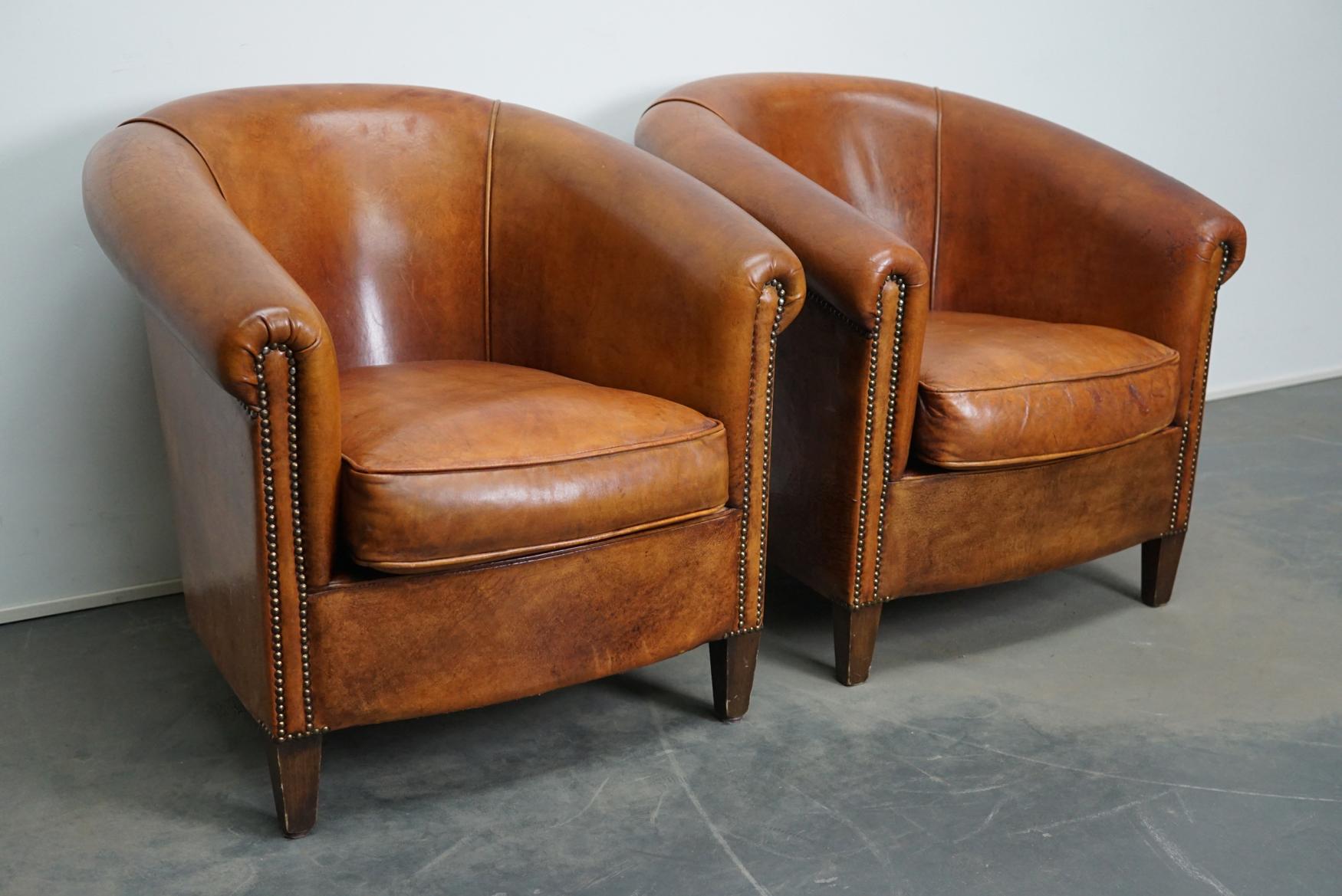 This pair of cognac-colored leather club chairs come from the Netherlands. They are upholstered with cognac-colored leather and feature metal rivets and wooden legs.