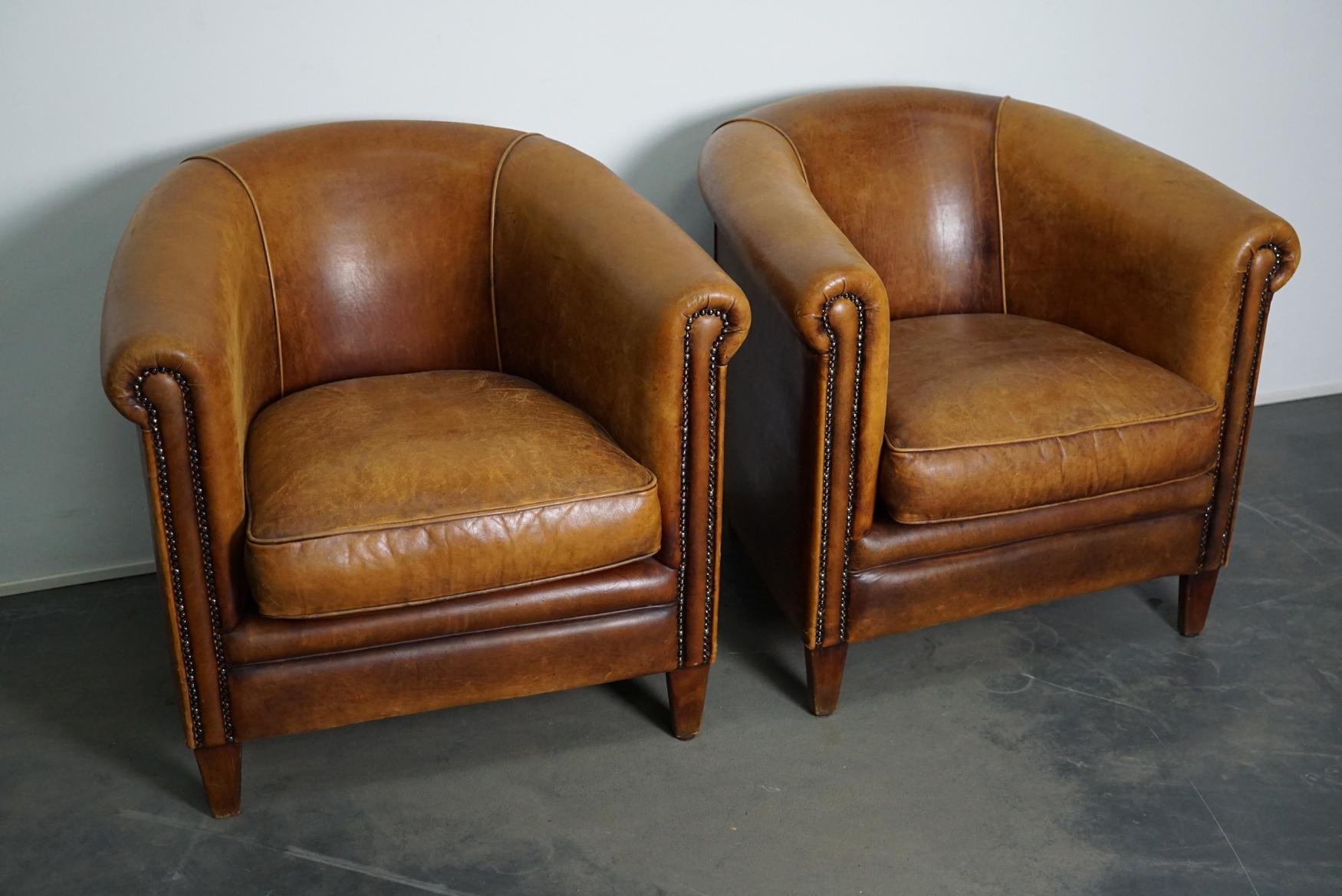 This pair of cognac-colored leather club chairs come from the Netherlands. They are upholstered with cognac-colored leather and feature metal rivets and wooden legs.