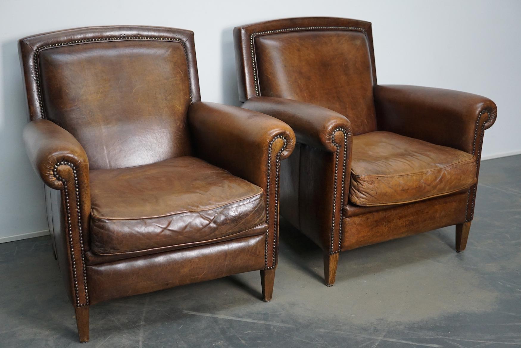 This pair of cognac-colored leather club chairs come from the Netherlands. They are upholstered with cognac-colored leather and feature metal rivets and wooden legs.