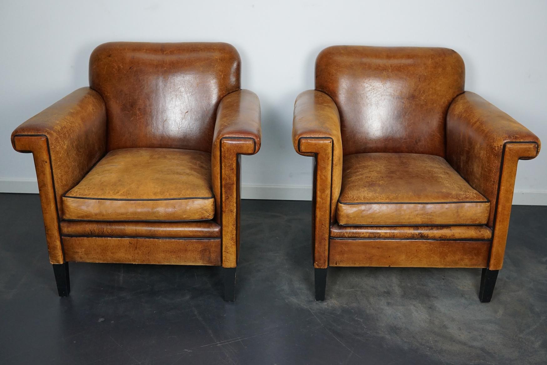 This pair of cognac-colored leather club chairs come from the Netherlands. They are upholstered with hand patinated leather and feature black piping and wooden legs.