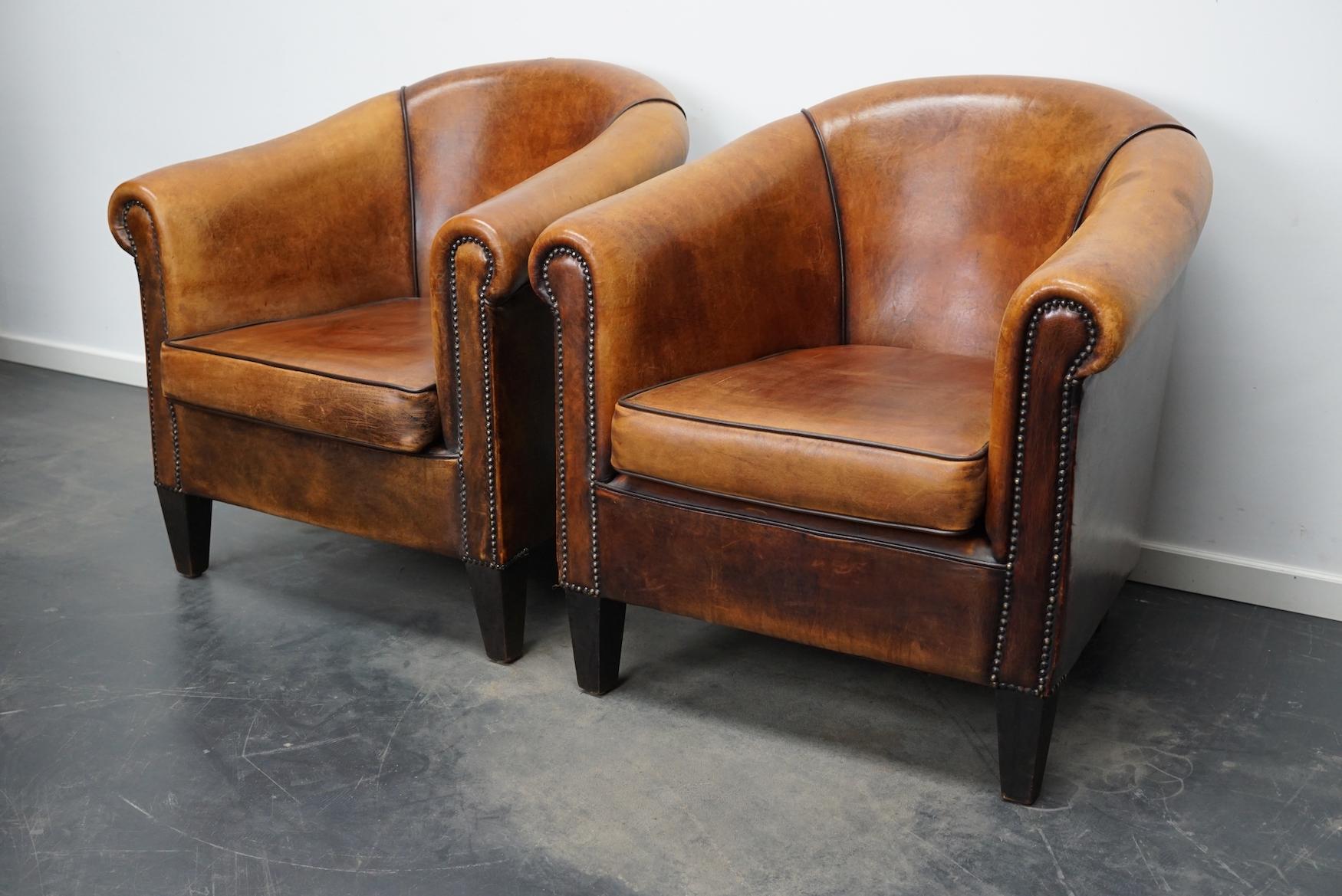 This pair of cognac-colored leather club chairs come from the Netherlands. They are upholstered with cognac-colored leather and feature metal rivets and wooden legs.