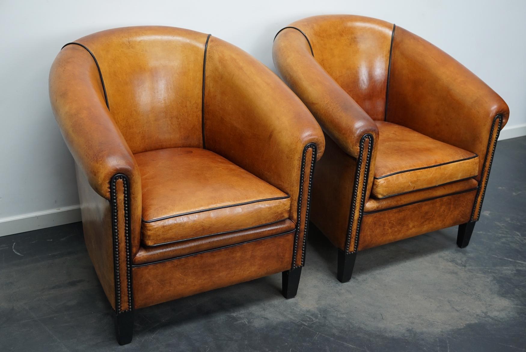This pair of cognac-colored leather club chairs come from the Netherlands. They are upholstered with hand patinated leather and feature black piping and wooden legs.