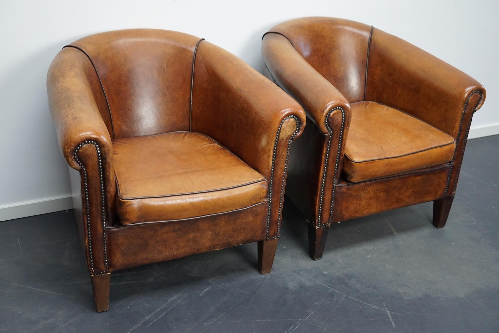 This pair of cognac-colored leather club chairs come from the Netherlands. They are upholstered with cognac-colored leather and feature metal rivets and wooden legs. This set also includes the footstool which is quite uncommon.