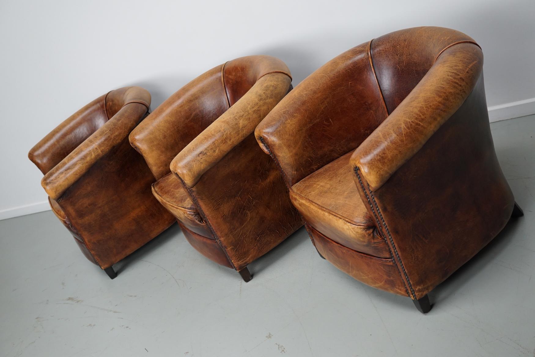 This set of three cognac-colored leather club chairs come from the Netherlands. They are upholstered with cognac-colored leather and feature metal rivets and wooden legs. 