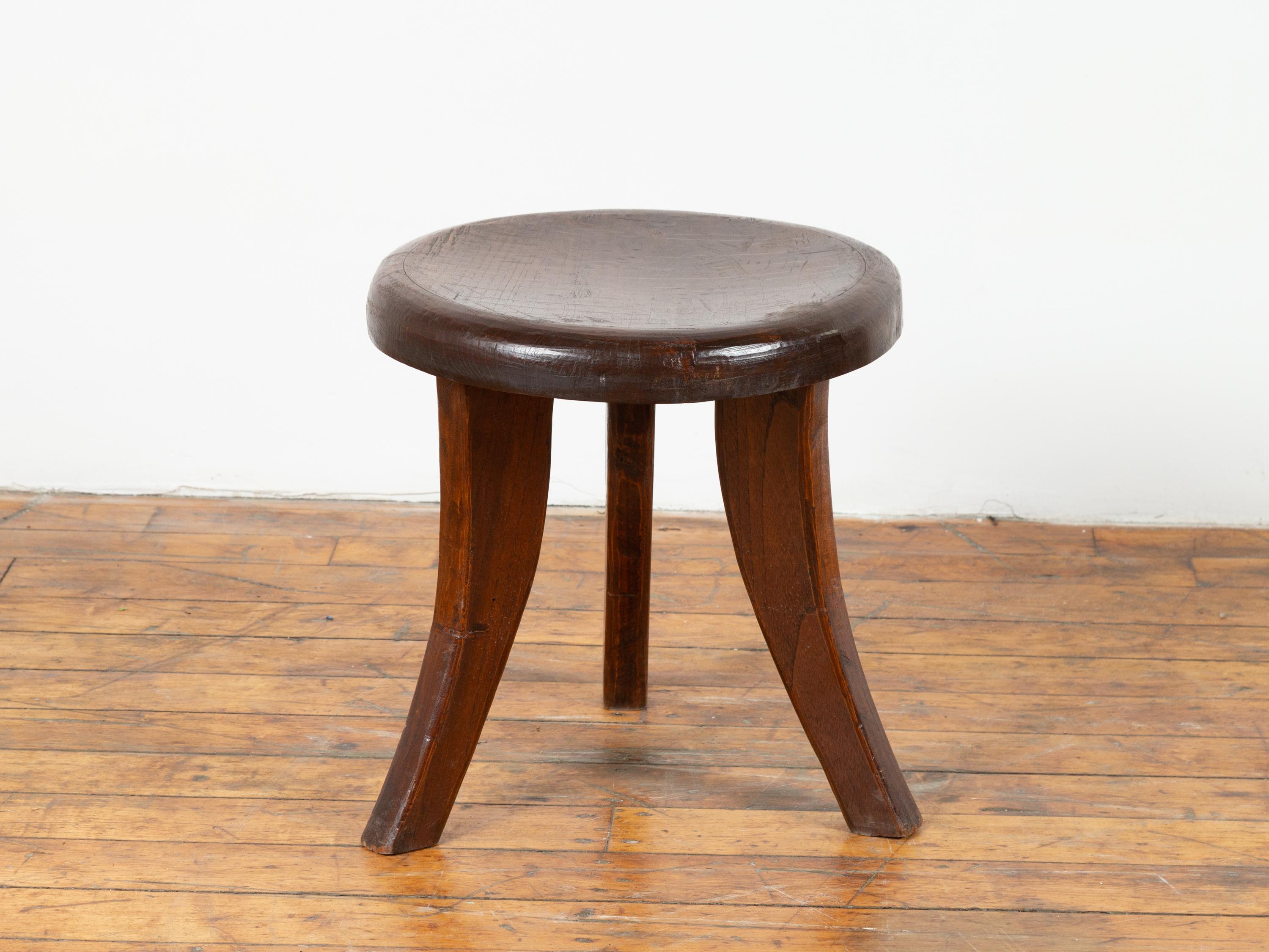 A vintage Dutch Colonial three-legged wooden stool. Born in Indonesian during the first half of the 20th century, this rustic stool features a circular top resting on three saber splaying legs. Simple and unpretentious, it will make for a great