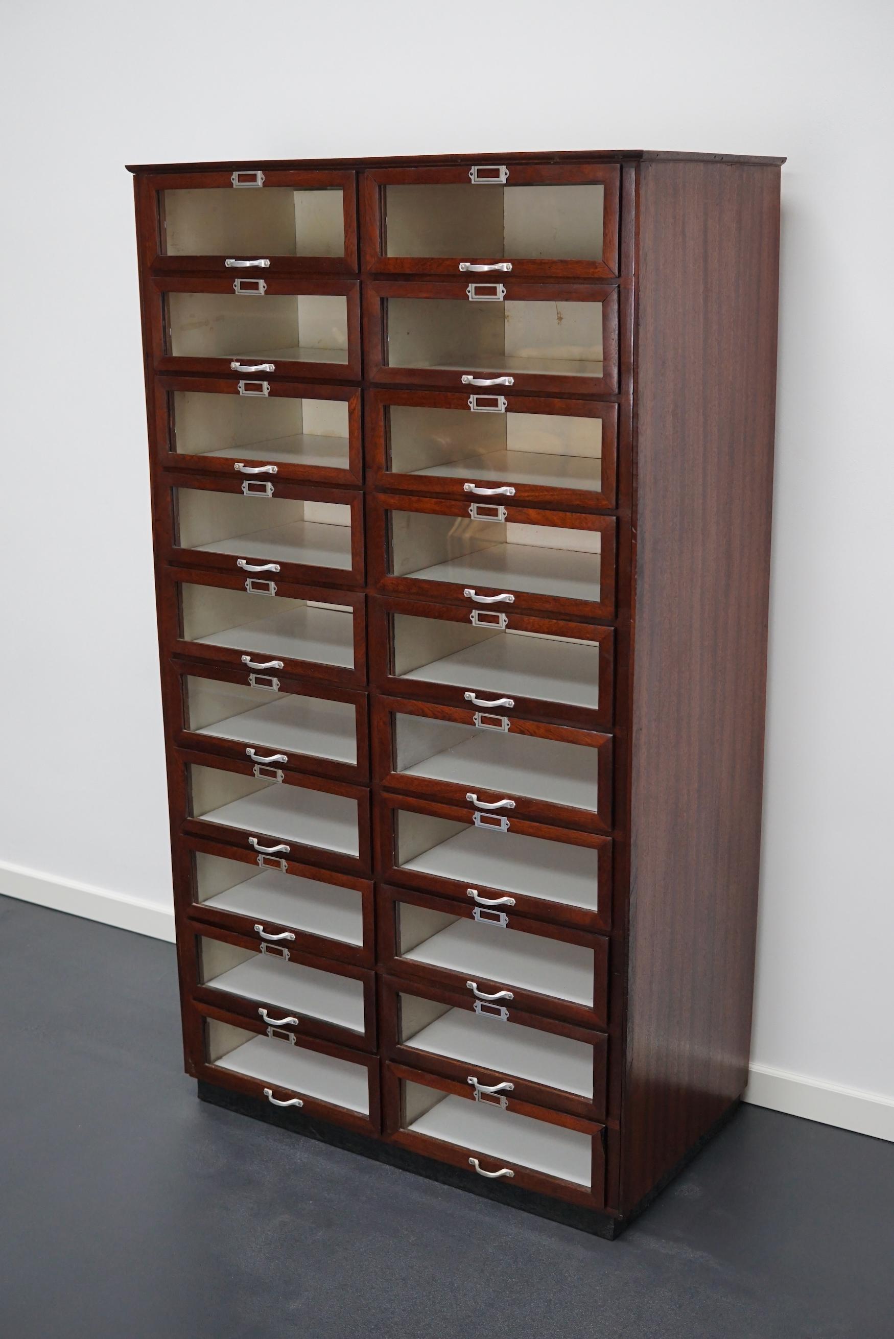 This haberdashery cabinet was produced during the 1930s in the Netherlands. This piece features 20 drawers in mahogany with glass fronts and metal handles. It was originally used in a shop for sewing supplies and fabrics in Amsterdam. The interior