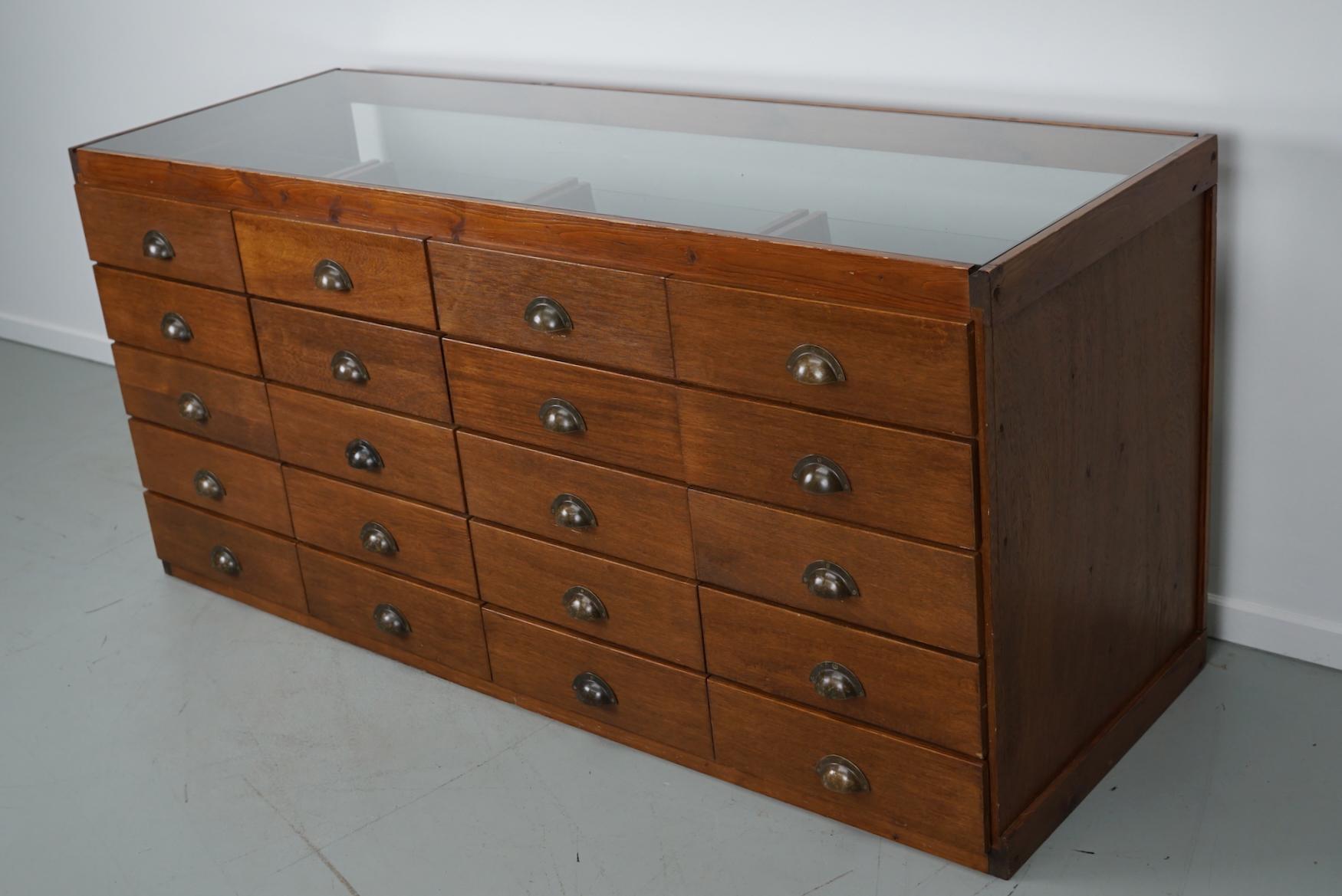 This vintage haberdashery shop counter dates from the 1960/70s and was handmade in the Netherlands. It features a wooden frame, glass casing and drawers in oak veneer with brass handles.