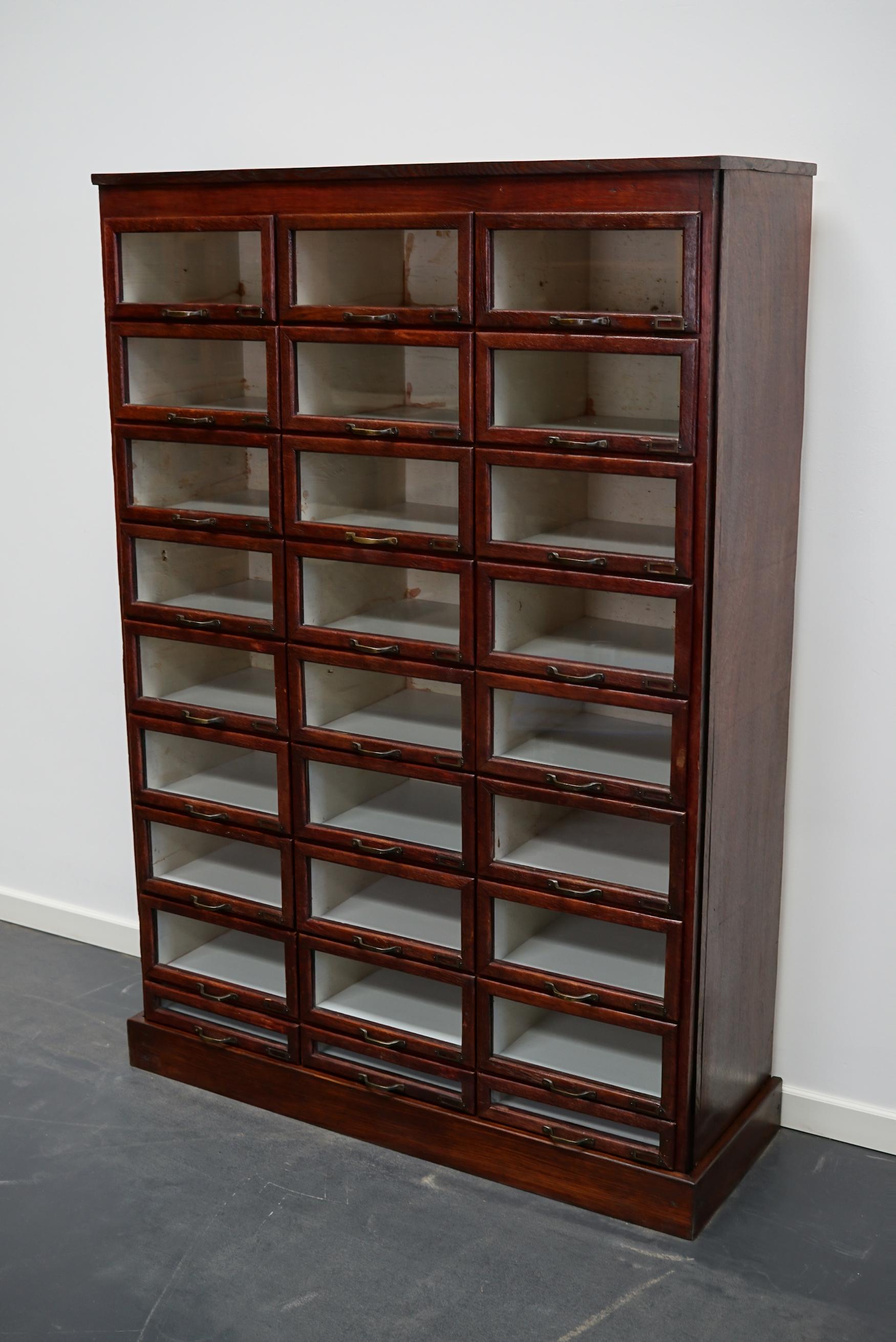 This haberdashery cabinet was produced during the 1930s in the Netherlands. This piece features 27 paper covered drawers in mahogany stained oak with glass fronts, brass handles and name card holders. It was originally used in a shop for sewing