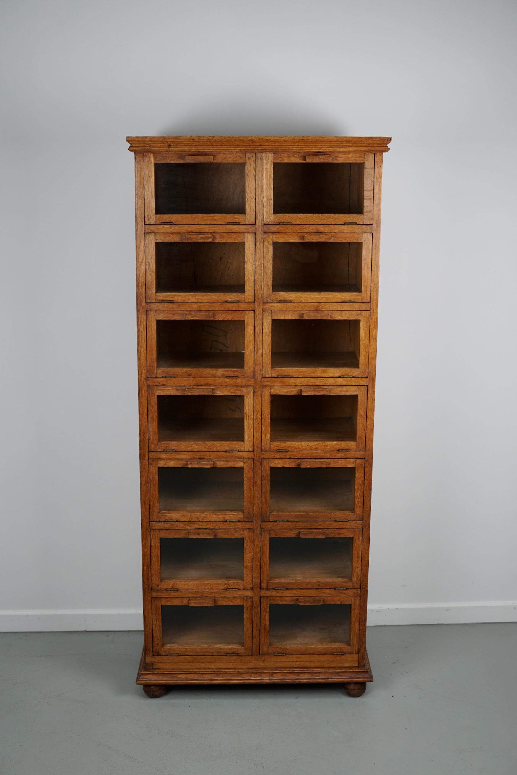 This haberdashery cabinet was produced during the 1930s in the Netherlands. It features 14 drop down doors in oak with glass fronts and oak handles. It was originally used in a warehouse for plant seeds in the city of Delft. The interior dimensions