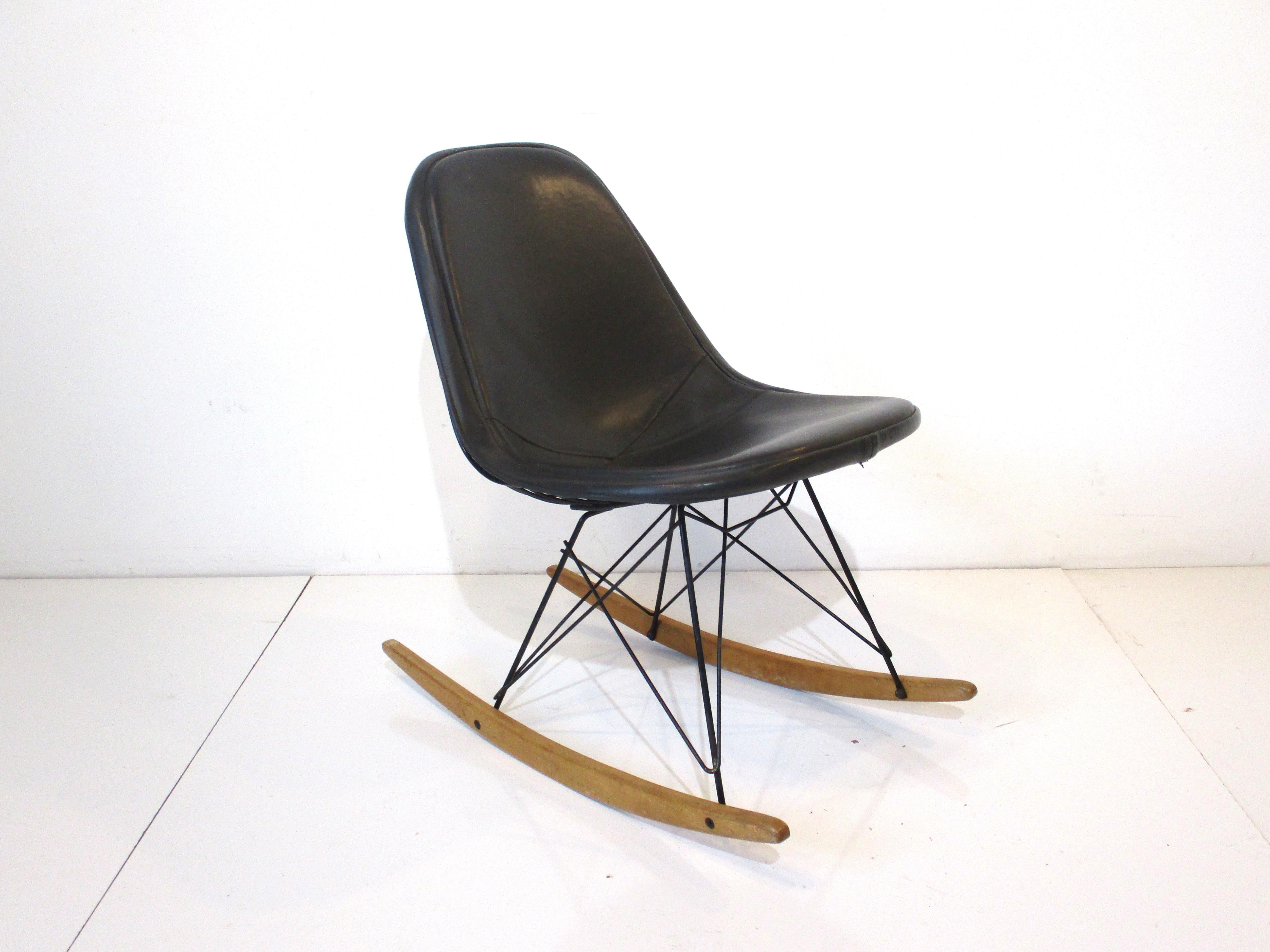 A rare satin black metal wire Eames rocking chair with birch wood runners and dark charcoal Naugahyde upholstered seat cover. The chair retains the fabric tag to the cover made by the Herman Miller furniture company Pasadena California.