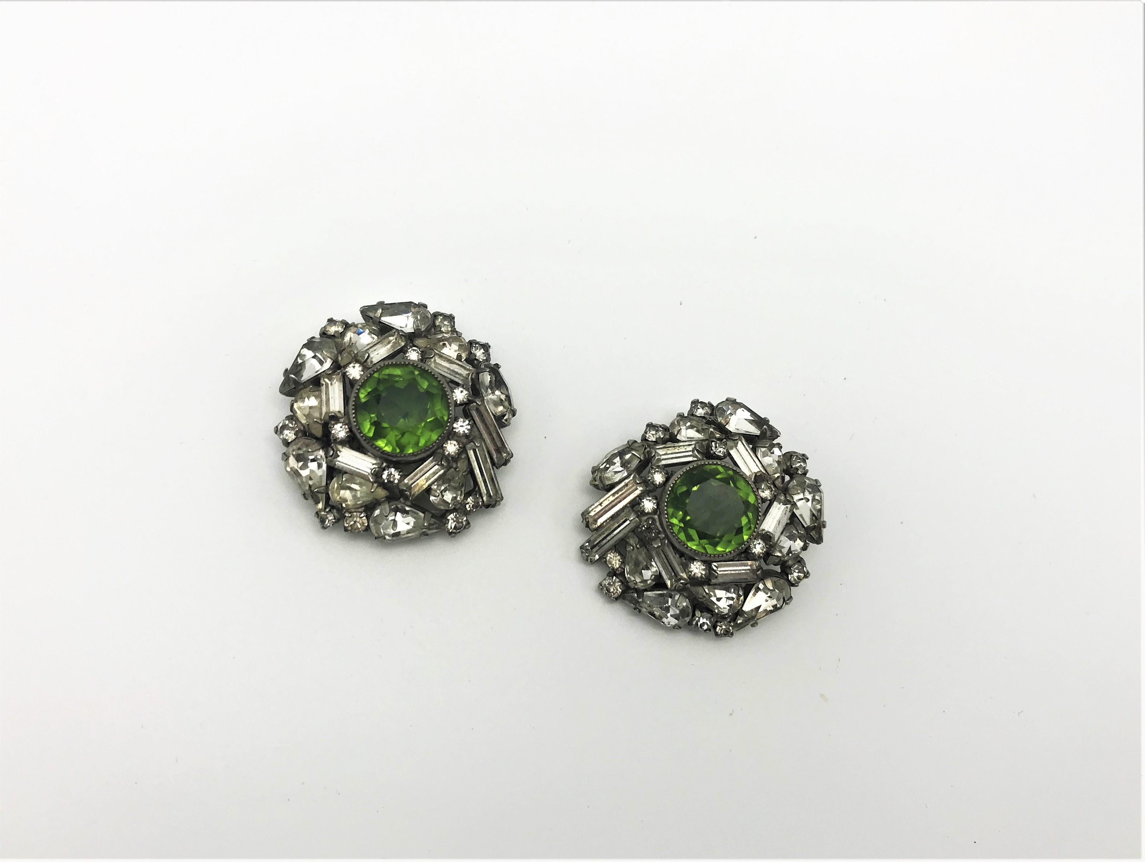 A pair of very nice and wearable ear clip with different shapes and cuts of rhinestones. In the middle there is a 1,5 cm large cut green rhinestone. He is made of silver metal and coms from the USA about 1940 years. 
Measurement: Diameter 3 cm, clip