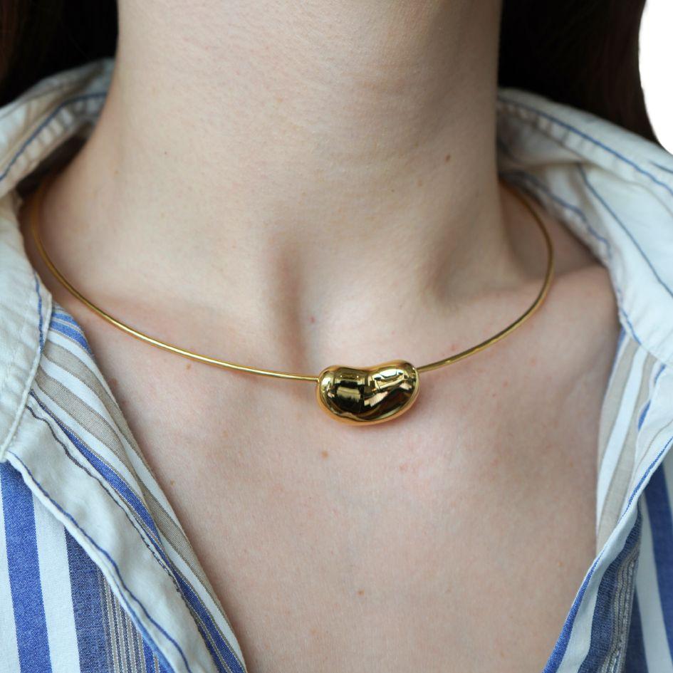 This vintage Tiffany & Co. necklace by Elsa Peretti features her iconic “Lucky Bean” design in 18k yellow gold. Crafted in the 1980s, the piece showcases a polished bean pendant strung on a minimalist 18k gold wire necklace, offering a sleek and