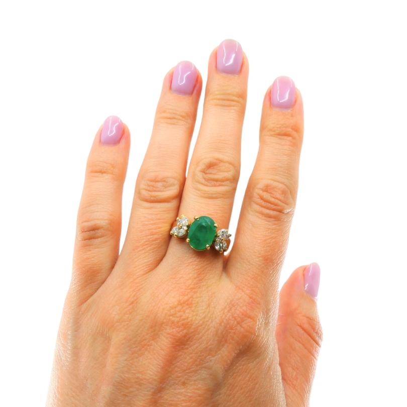 14k yellow gold ring featuring an emerald as the center stone, flanked by diamonds. The emerald is oval-shaped and a vibrant green, while the diamonds arranged in a floral  motif on either side of the emerald, adding sparkle to the design.  The ring
