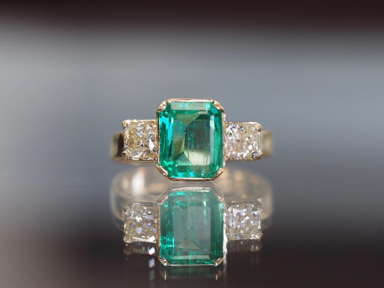 This beautiful vintage ring 18 karat yellow gold ring includes a natural emerald center weighing 2.13ct center measuring 8.80mm x 7.33mm. It is accented with one radiant diamond on each side of the center weighing 0.73 carat total weight measuring