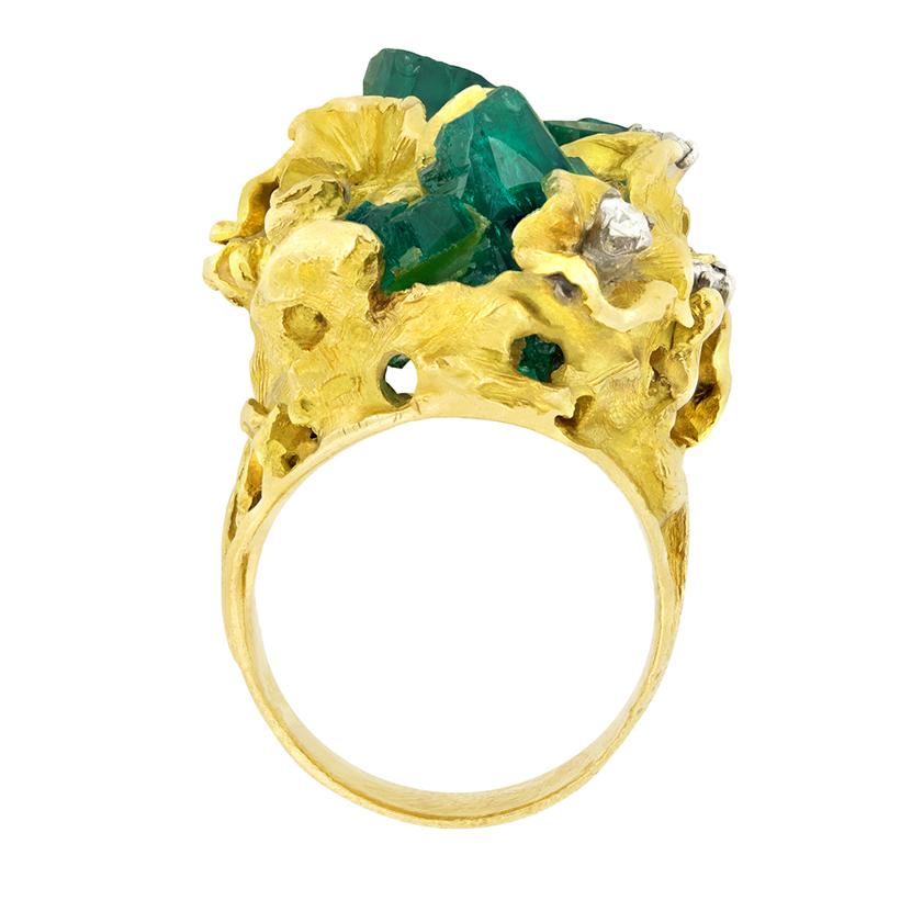 Dieser in den 1960er Jahren gefertigte Blickfang ist aus 18 Karat Gelbgold gefertigt. Aus einem fließenden Kopf aus Rohgold blühen eine Reihe von Smaragdclustern im Rohschliff, die von fünf runden Brillanten hervorgehoben werden. Die Diamanten haben